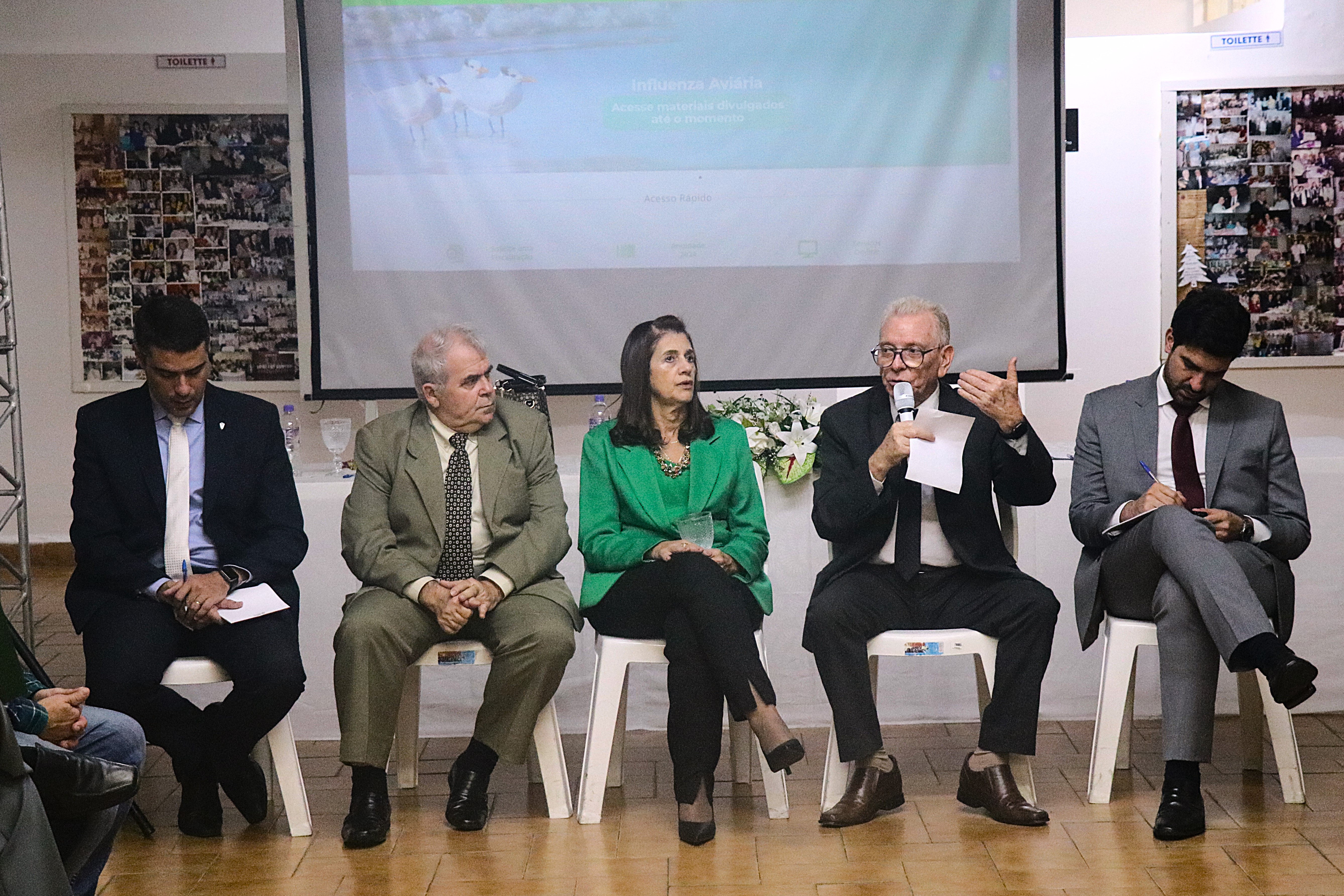 Quatro pessoas, três homens e uma mulher, sentados em cadeiras lado a lado, ouvem um deles ao microfone.