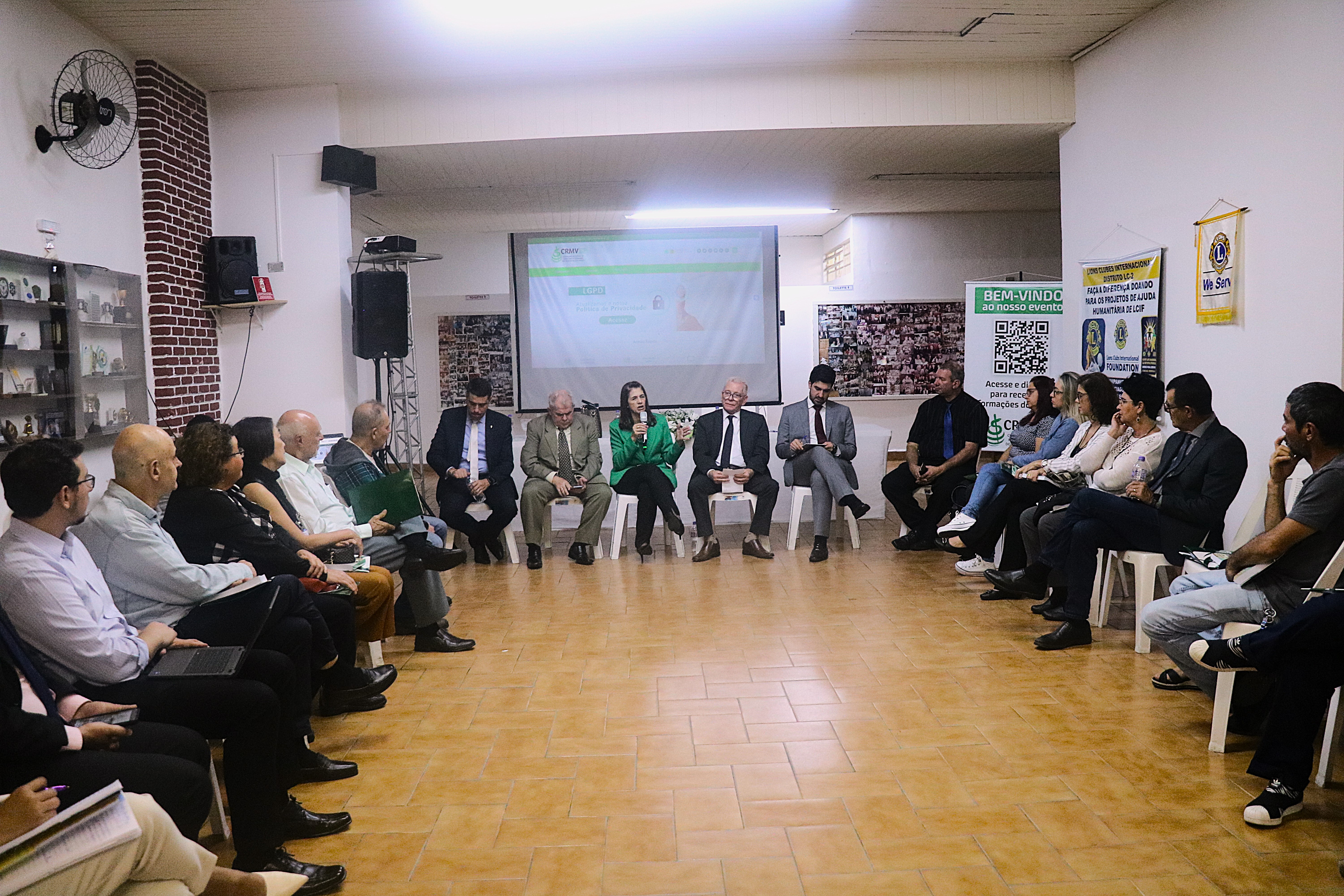 Homens e mulheres, sentados em cadeiras, organizadas em formato de U, ouvem a tesoureira do CRMV-SP falar ao microfone no auditório do evento.
