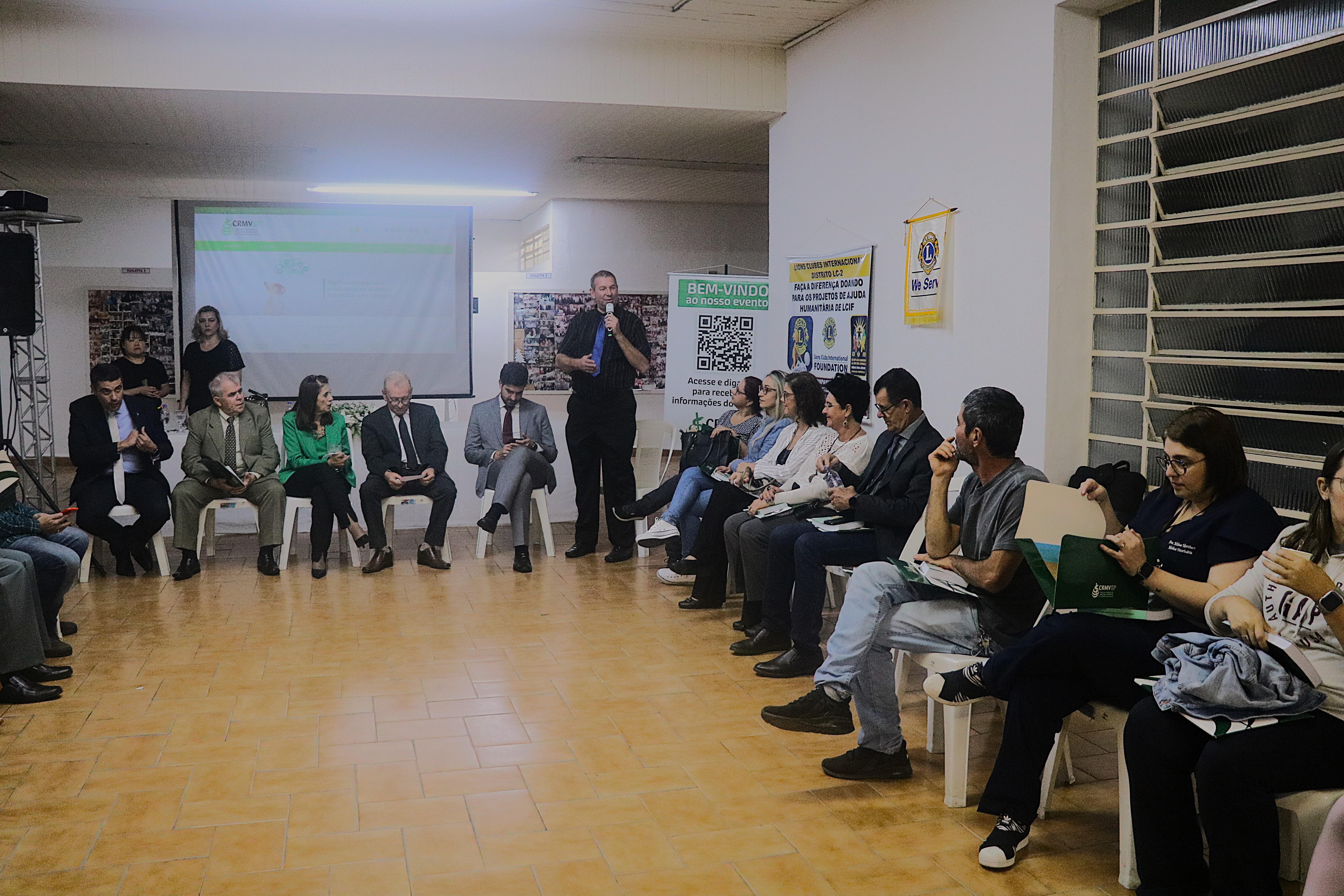 O presidente da Sociedade dos Médicos-veterinários da Zona Norte de São Paulo, em pé, fala ao microfone para a plateia, sentada em cadeiras, organizadas em formato de U.