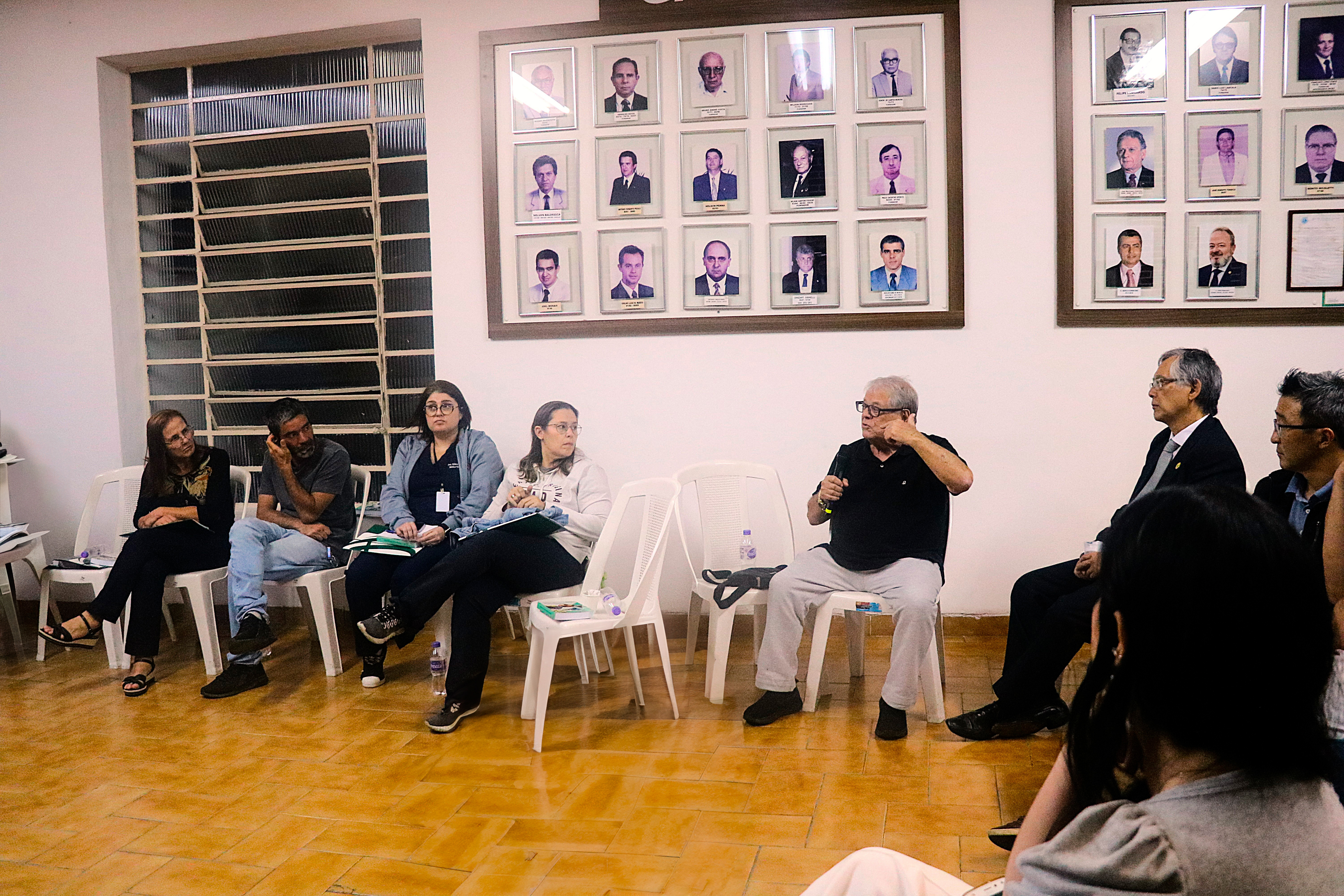 Homens e mulheres, sentados em cadeiras, lado a lado, ouvem um dos participantes falar ao microfone no auditório do evento.