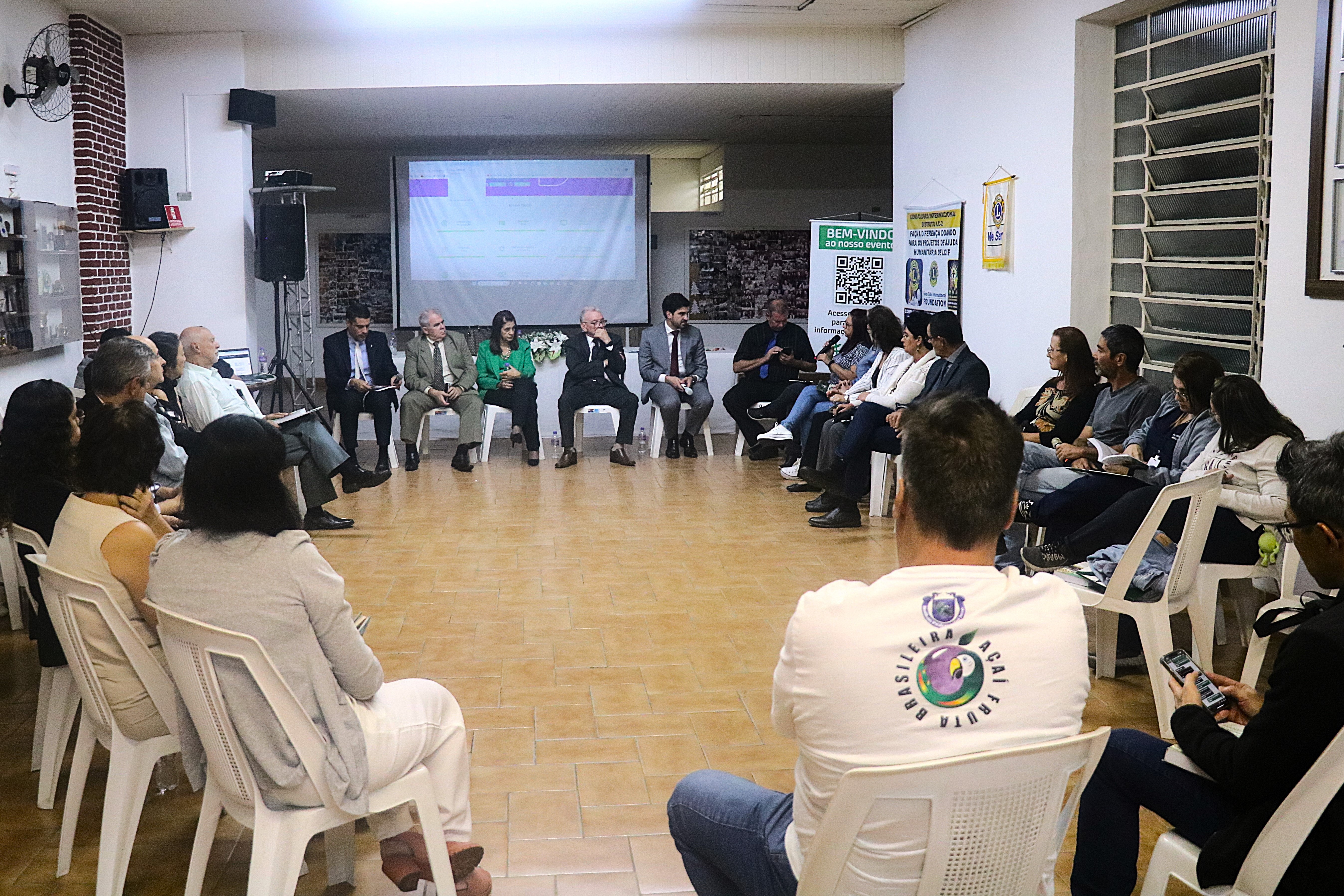Homens e mulheres, sentados em cadeiras, organizadas em formato de U, ouvem uma das participantes do evento falar ao microfone no auditório do evento.