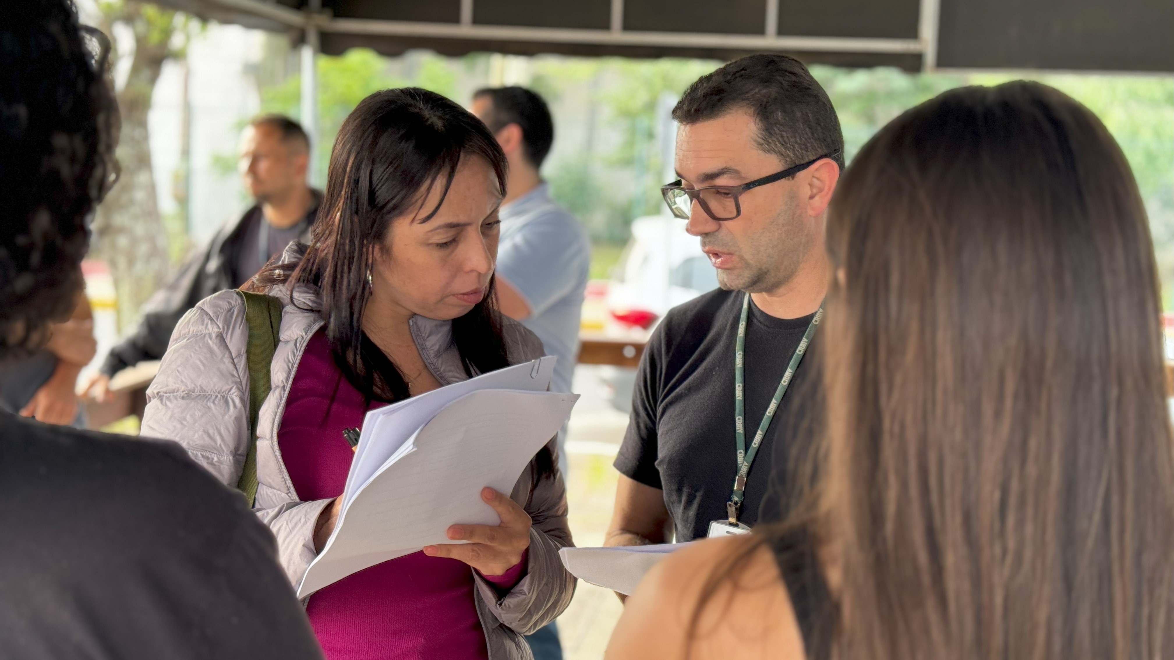 Mulher vestindo blusa rosa e casaco cinza segurando canetas e papéis conversa com homem vestindo blusa preta e usando óculos marrom. Há duas pessoas de costas também na imagem.