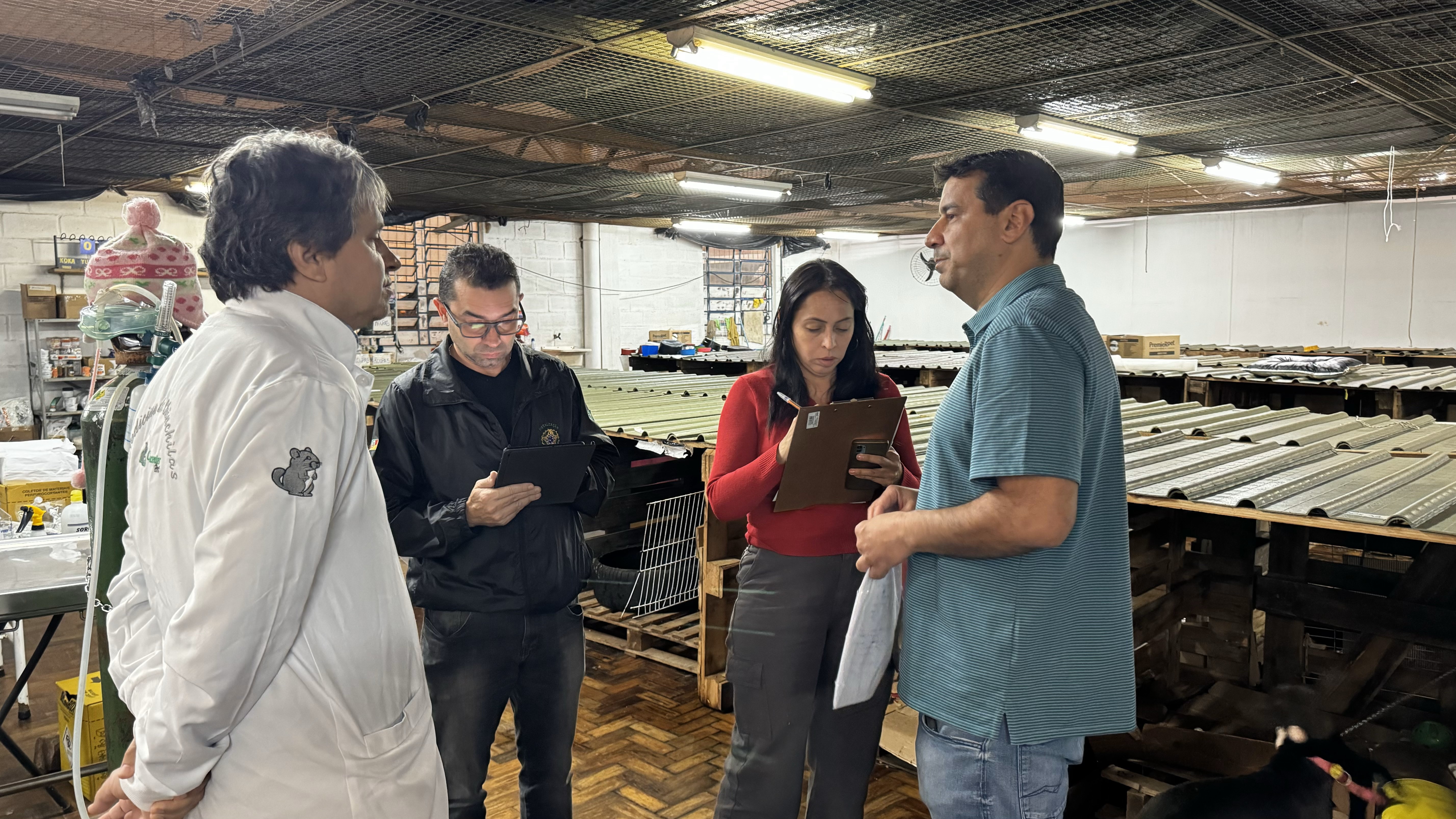 Médico-veterinário, fiscal sanitário, mulher escrevendo em prancheta e homem usando blusa azul e calça jeans fotografados em recinto de animais no Rio Grande do Sul.