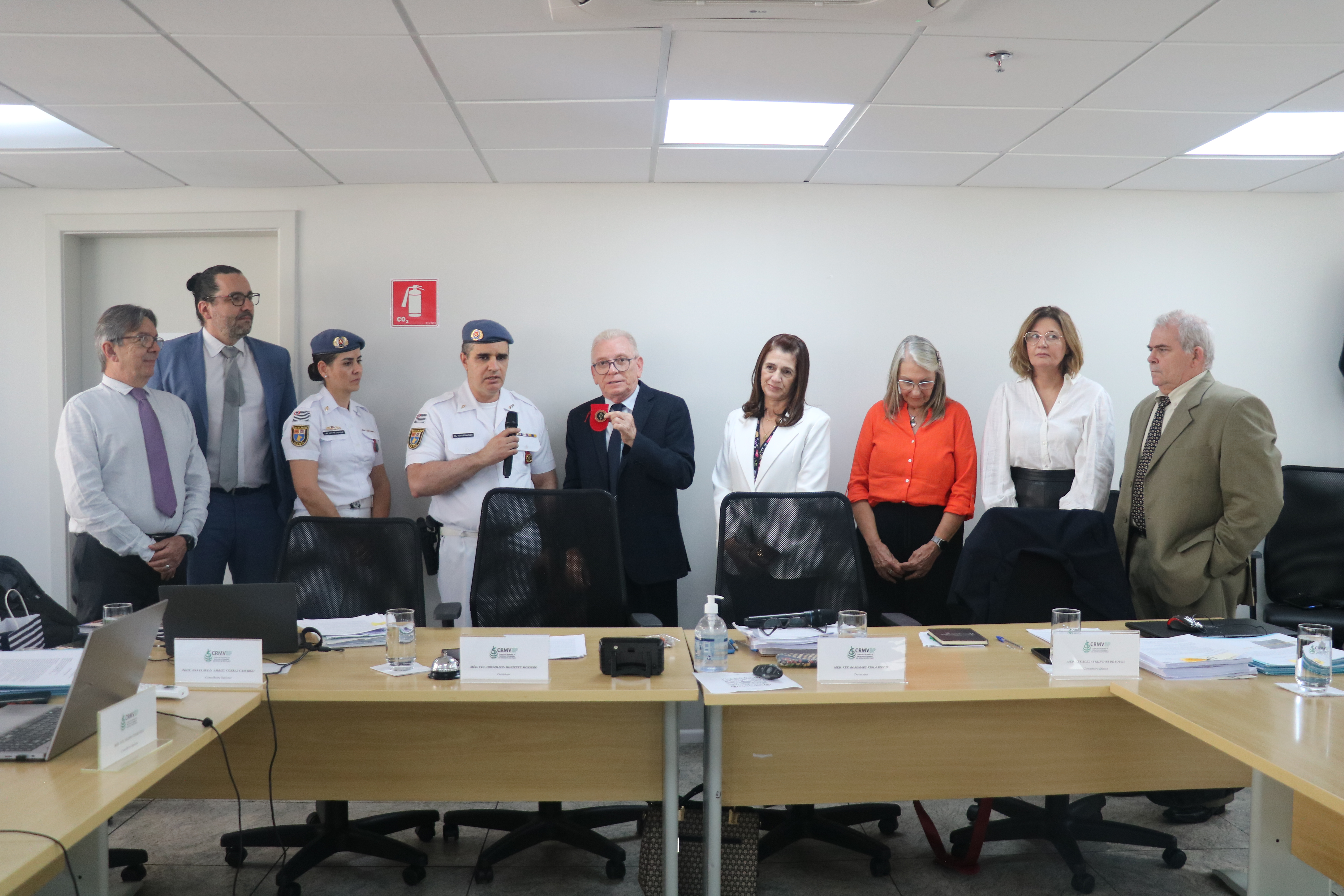 Nove pessoas em pé na sala da plenária, no centro da foto, médicos-veterinários(uma mulher e um homem) militares em pé recebendo medalha de homenagem das mãos do presidente do CRMV-SP.