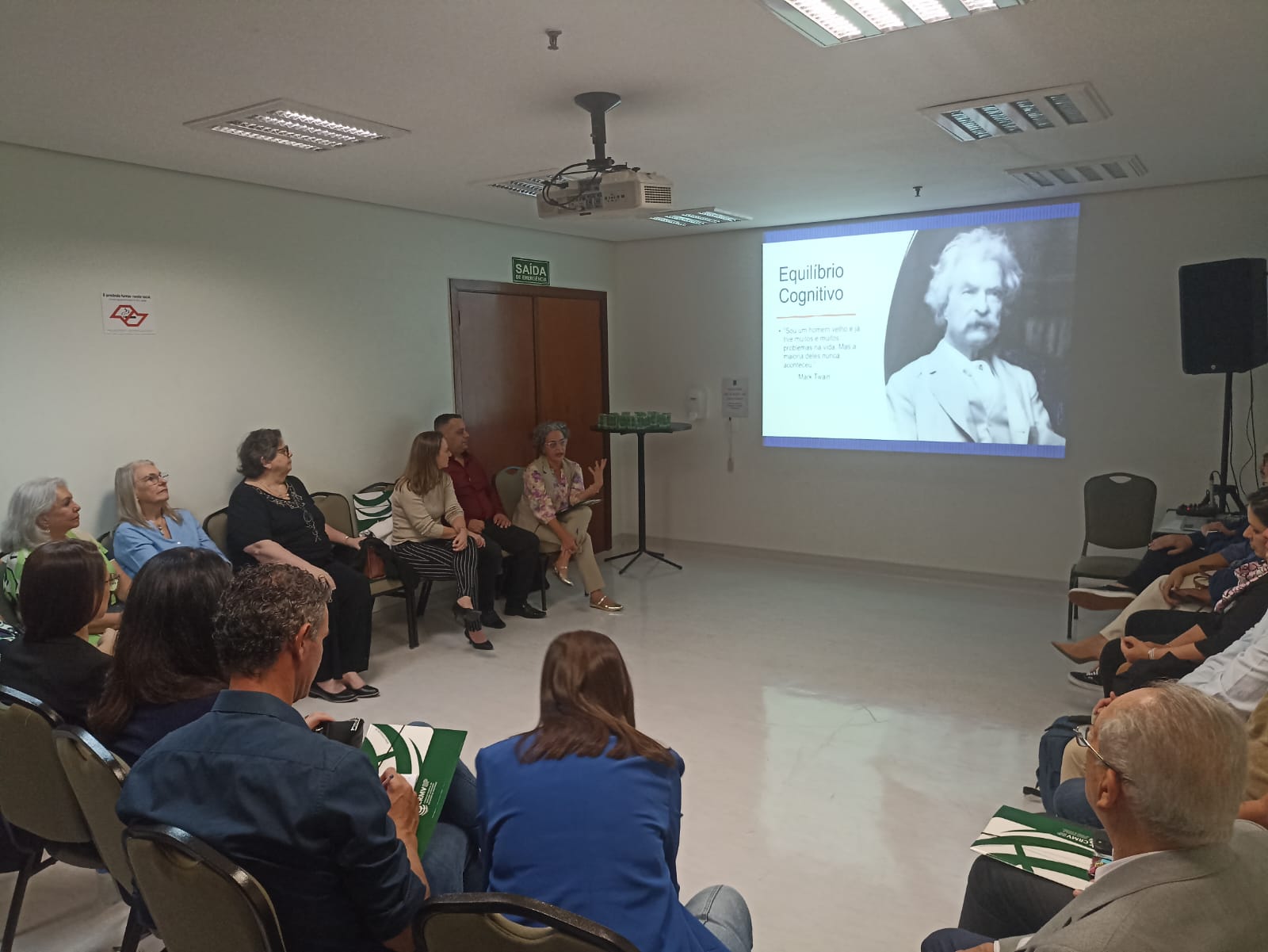 Pessoas sentadas em semicírculo durante reunião do Grupo de Apoio Mútuo(GAM) olhando apresentação na parede.