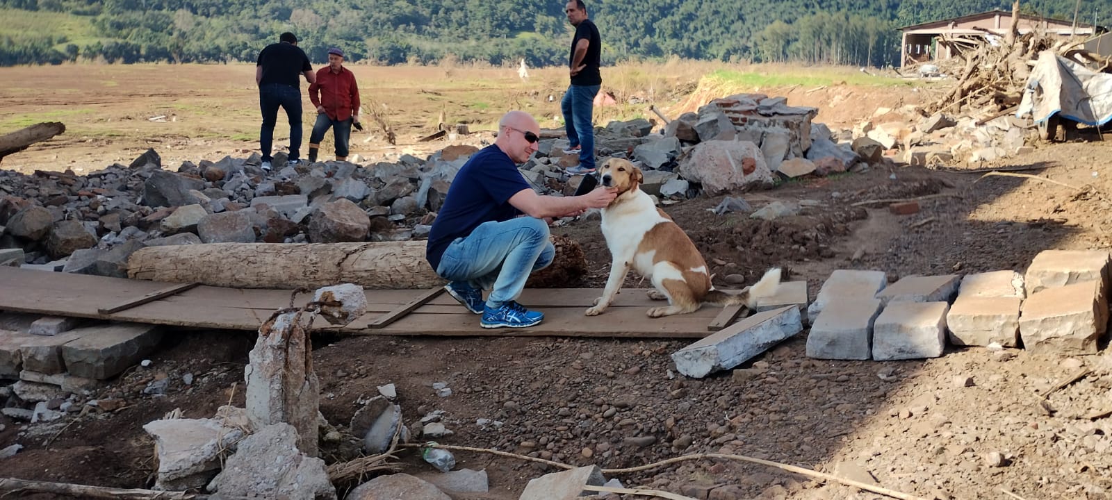 Homem branco usando tênis, calça jeans e blusa azul, faz carinho em cachorro adulto sem raça definida branco e marrom em local com escombros no Rio Grande do Sul.