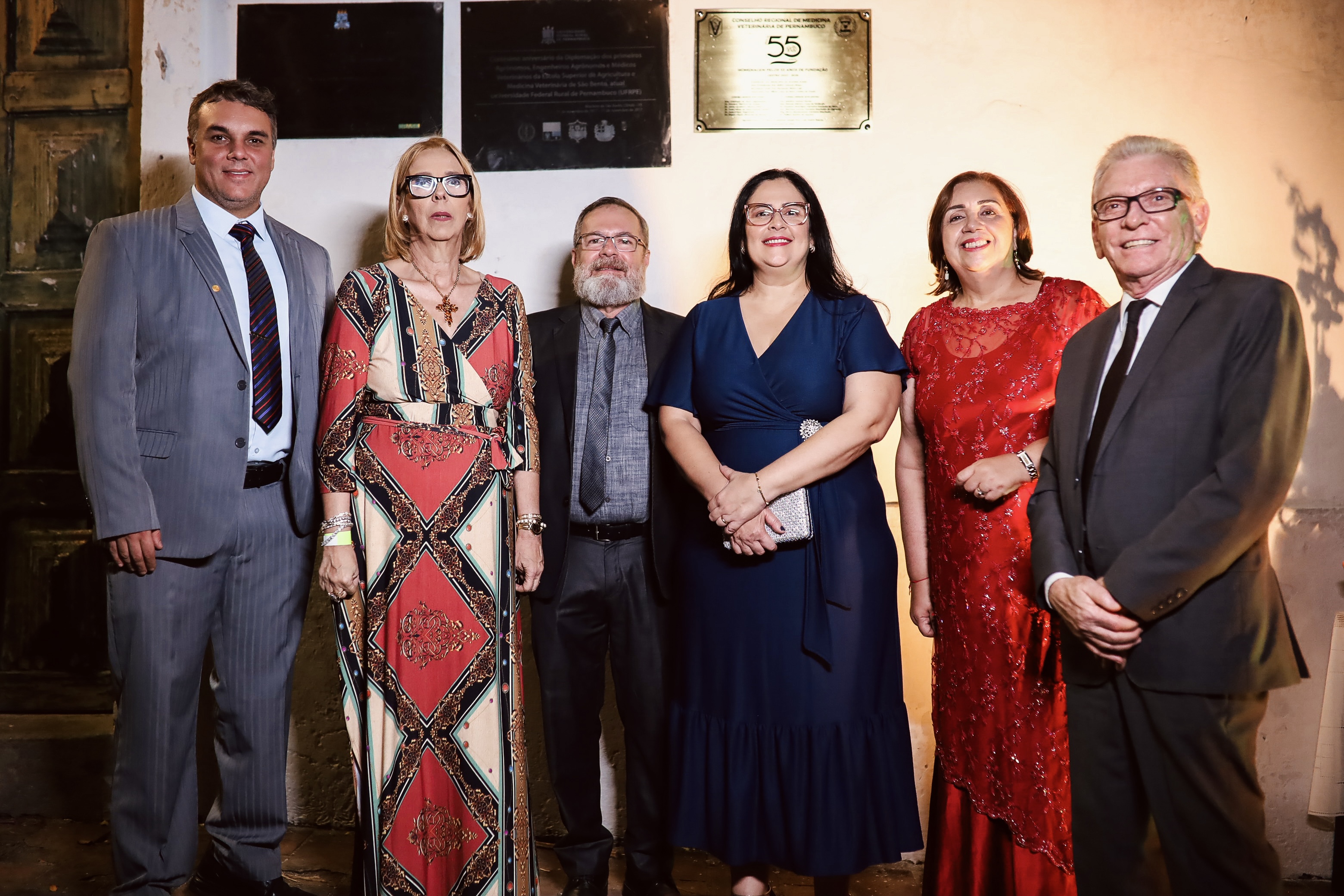 três homens e três mulheres, as mulheres de vestido de festa e os homens de terno e gravata, pousando sorridentes para foto.