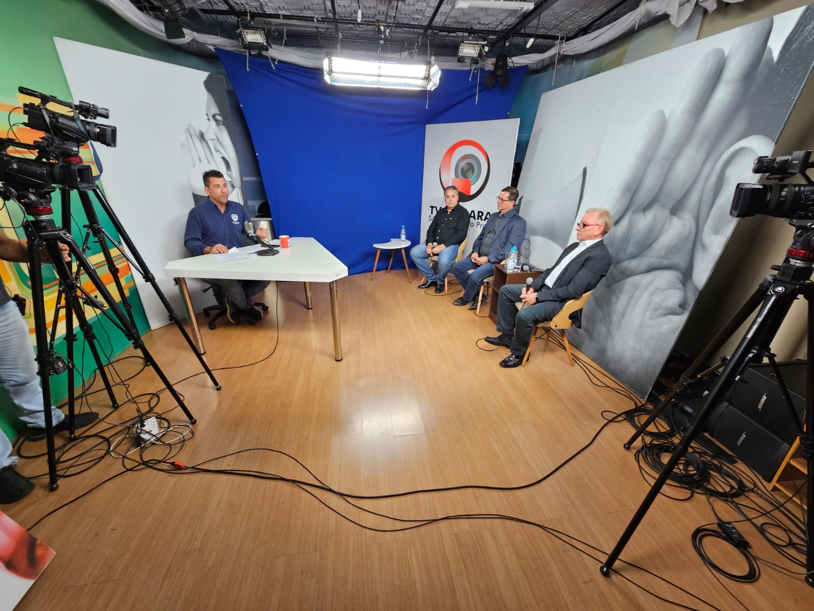 Em um estúdio de televisão, o apresentador está sentado à mesa, olhando para a câmera, com três homens, que serão entrevistados, sentados à sua frente.
