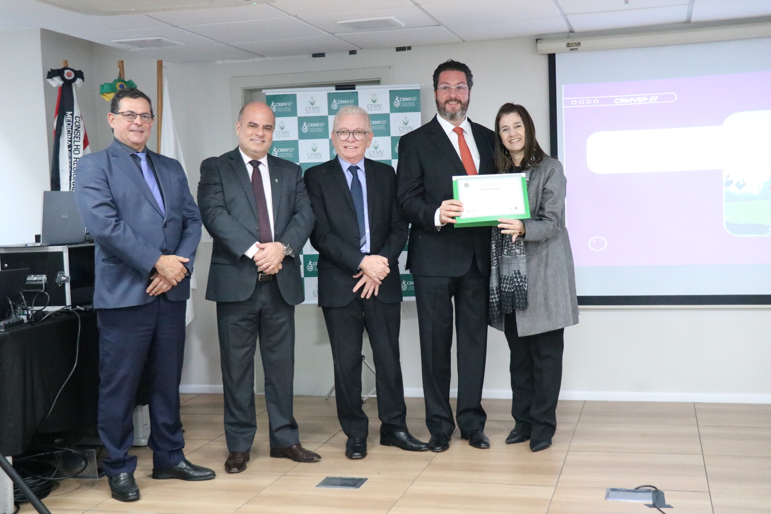 No palco, os membros da Diretoria Executiva do CRMV-SP posam ao lado de um presidente de Grupo de Trabalho homenageado com certificado em reconhecimento ao trabalho desempenhado.