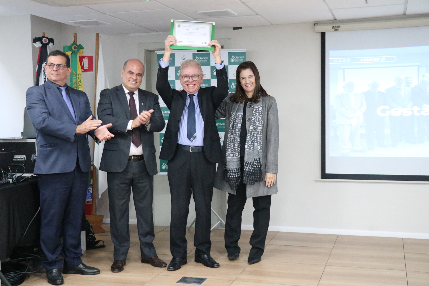 Ao lado dos demais membros da Diretoria Executiva, o presidente do CRMV-SP levanta as mãos para o alto mostrando o certificado que recebeu em reconhecimento pelos trabalhos desempenhados na função.