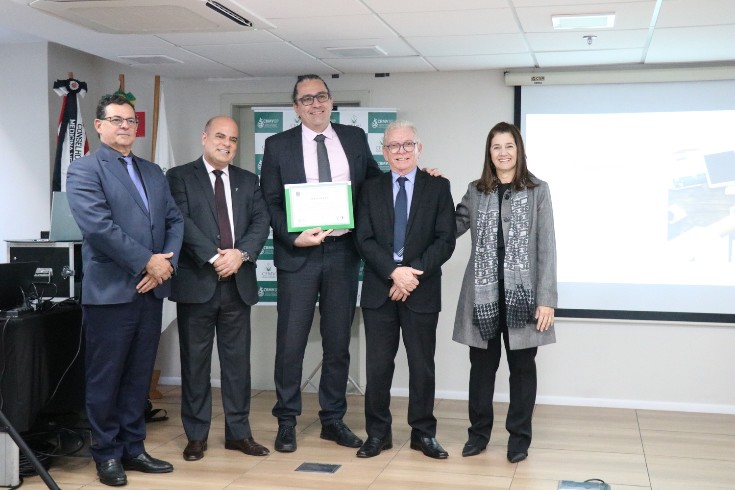 No palco, os membros da Diretoria Executiva do CRMV-SP posam ao lado de um presidente de Comissão Técnica homenageado com certificado em reconhecimento ao trabalho desempenhado.