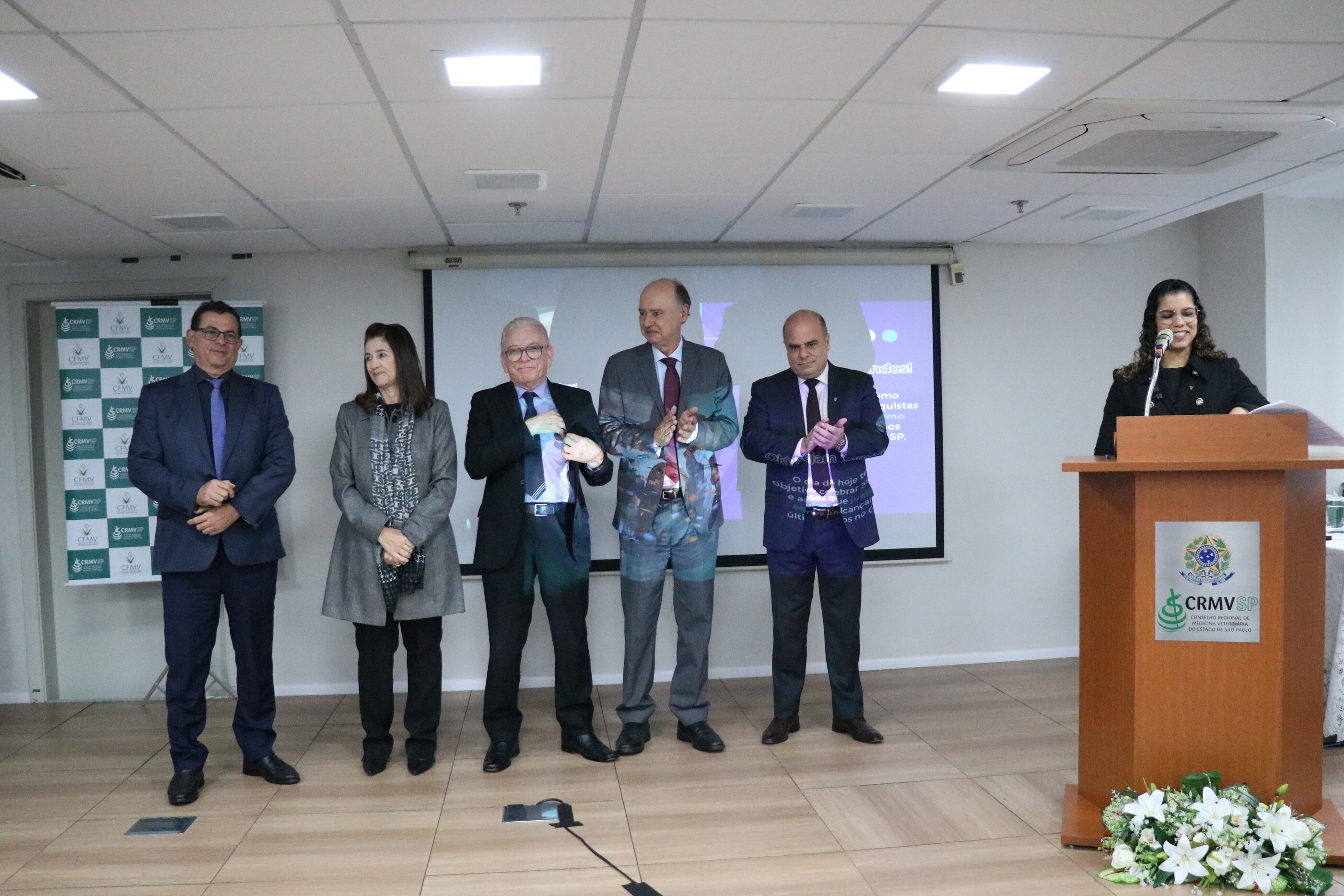 No palco do auditório, sei pessoas, duas mulheres e quatro homens. Uma das mulheres está no púlpito apresentando os demais, que estão em pé, lado a lado, para o início da cerimônia.