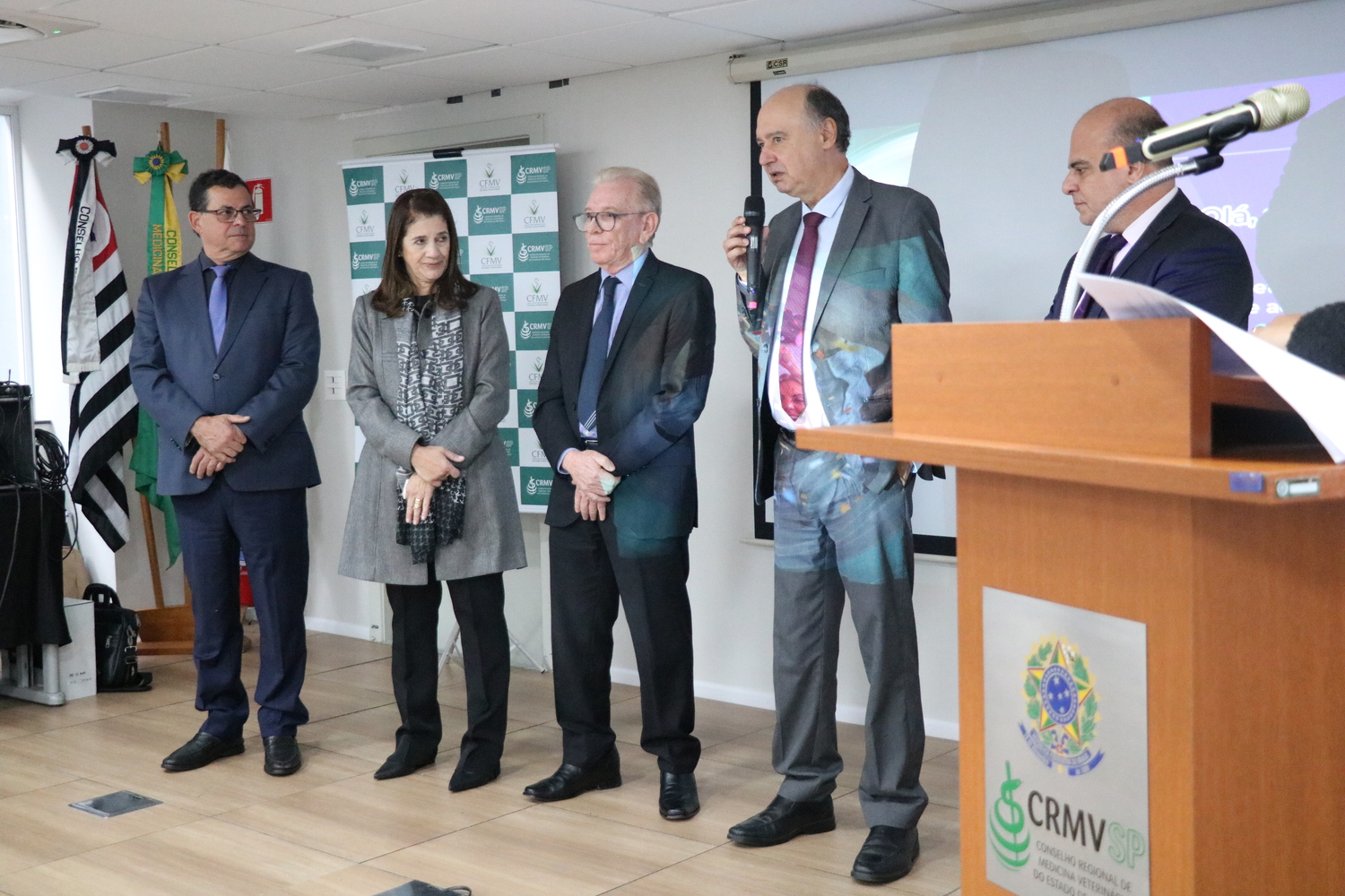 No palco do auditório, cinco pessoas sendo quatro homens e uma mulher, lado a lado, um dos homens fala ao microfone.