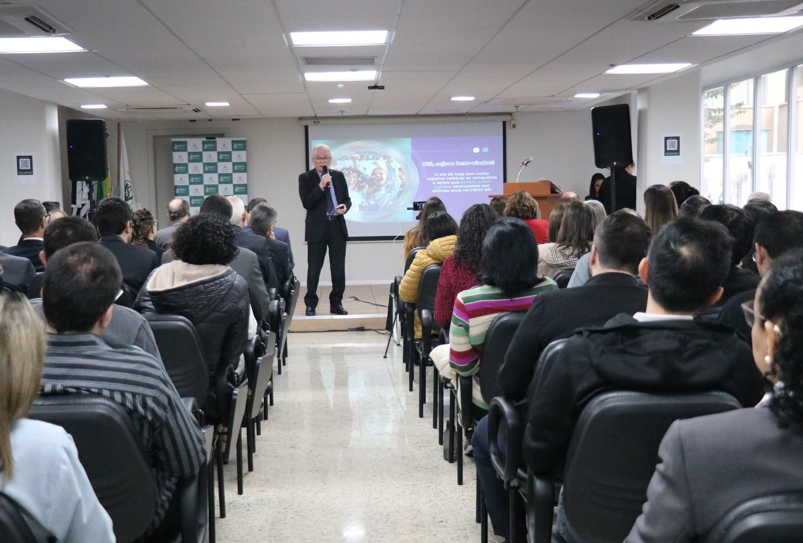 O presidente do CRMV-SP fala ao microfone, no palco, observado pela plateia, formada por homens e mulheres.