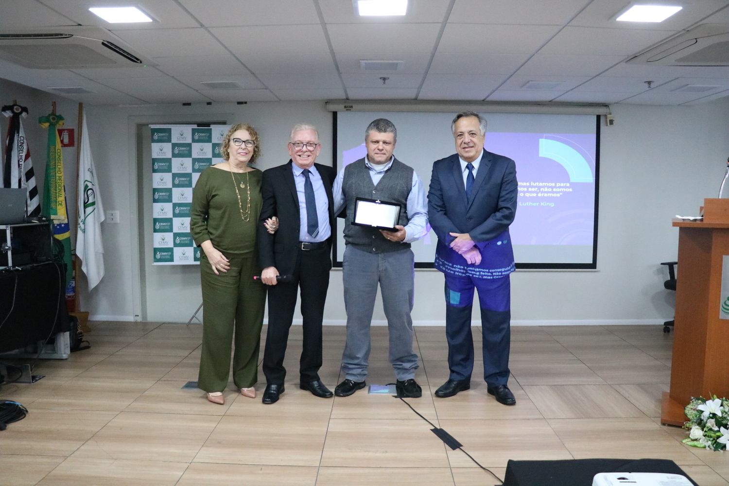 No palco do auditório, uma mulher e três homens, lado a lado. Um dos homens está sendo homenageado e segura uma placa.