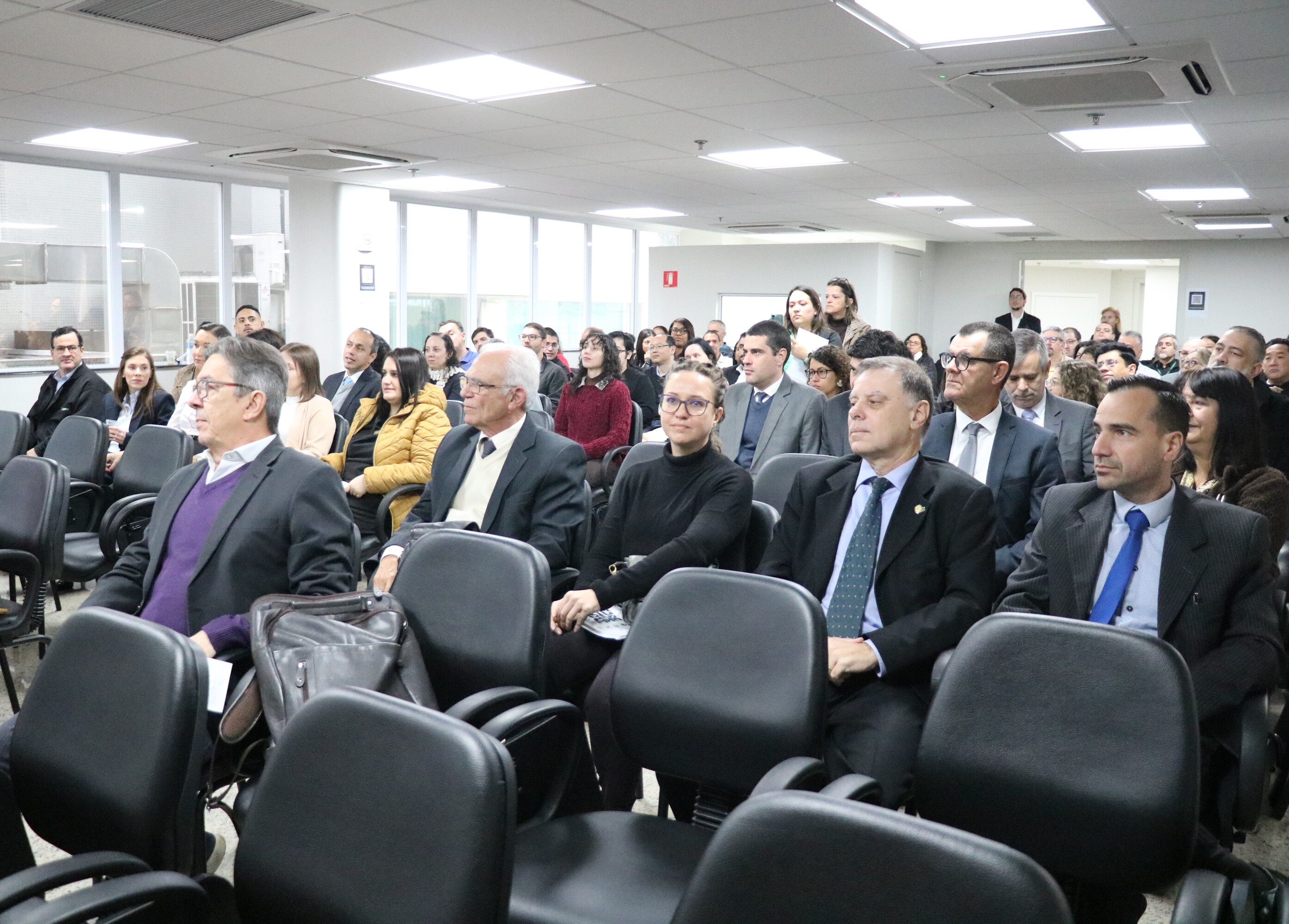 A plateia formada de homens e mulheres, sentados em cadeiras, assistindo ao evento.