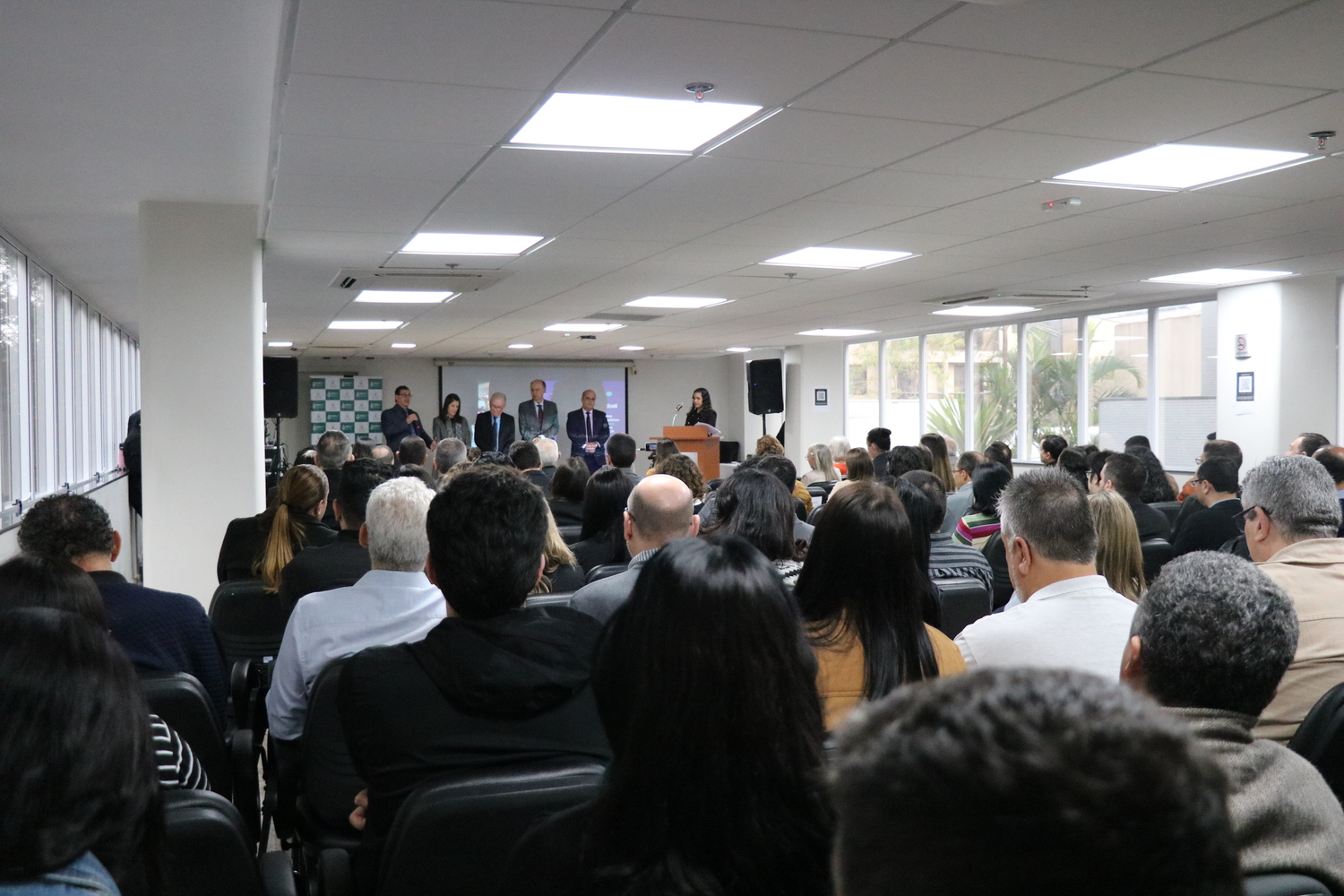 Imagem da plateia lotada e ao fundo o palco com seis pessoas, quatro homens e duas mulheres, sendo que uma delas apresenta o evento do púlpito.