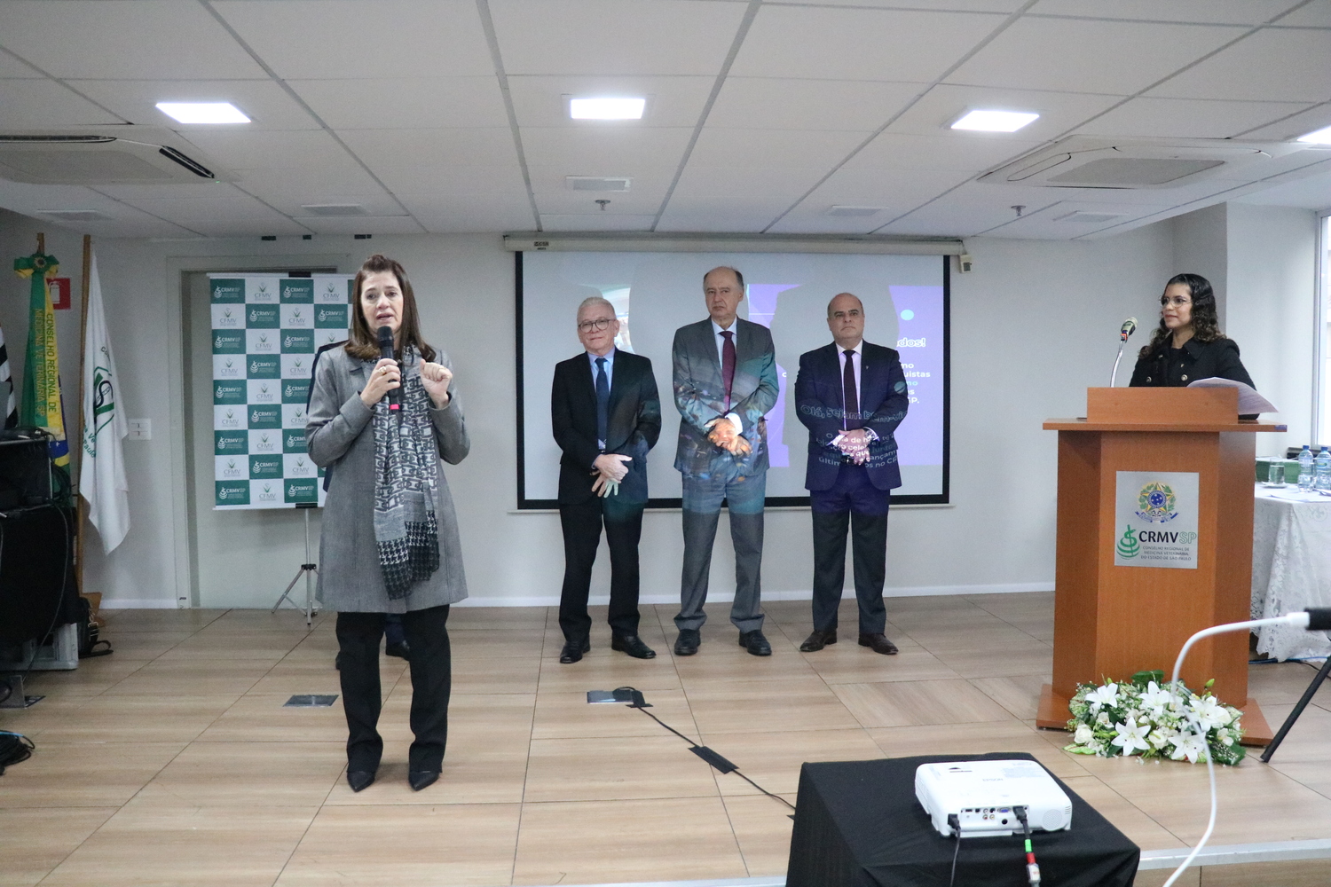 No palco do auditório, uma mulher fala ao microfone, observada por três homens e outra mulher, que apresenta o evento, posicionada no púlpito.