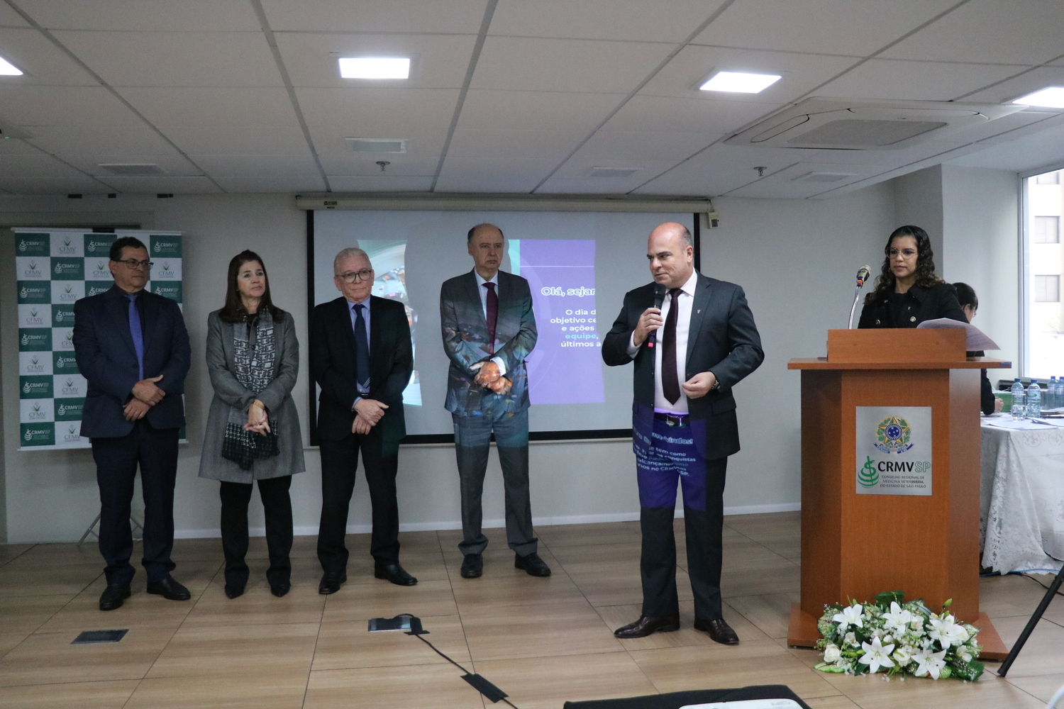 No palco do auditório, um homem fala ao microfone, observado por três homens e uma mulher ao fundo, e outra mulher, que apresenta o evento, posicionada no púlpito.
