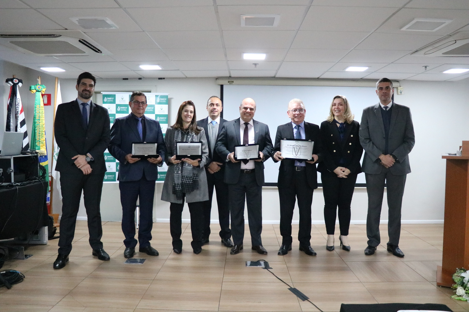 No palco, oito pessoas, entre mulheres e homens, sendo que os membros da Diretoria Executiva do CRMV-SP, posam com suas placas, ao lado dos funcionários que as entregaram.