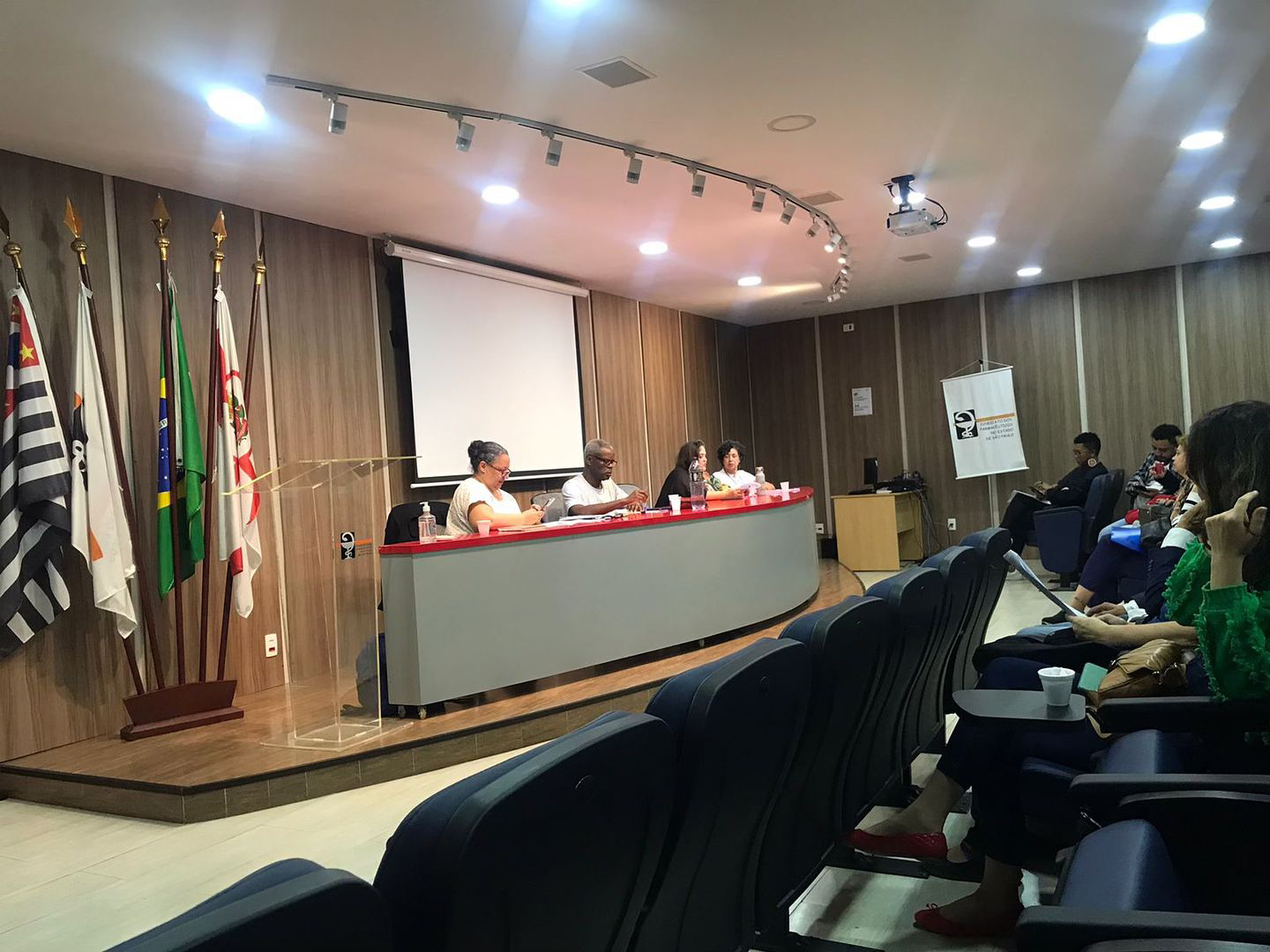 Quatro pessoas sentadas na mesa principal palestrando em um auditório em reunião do FCAFs.