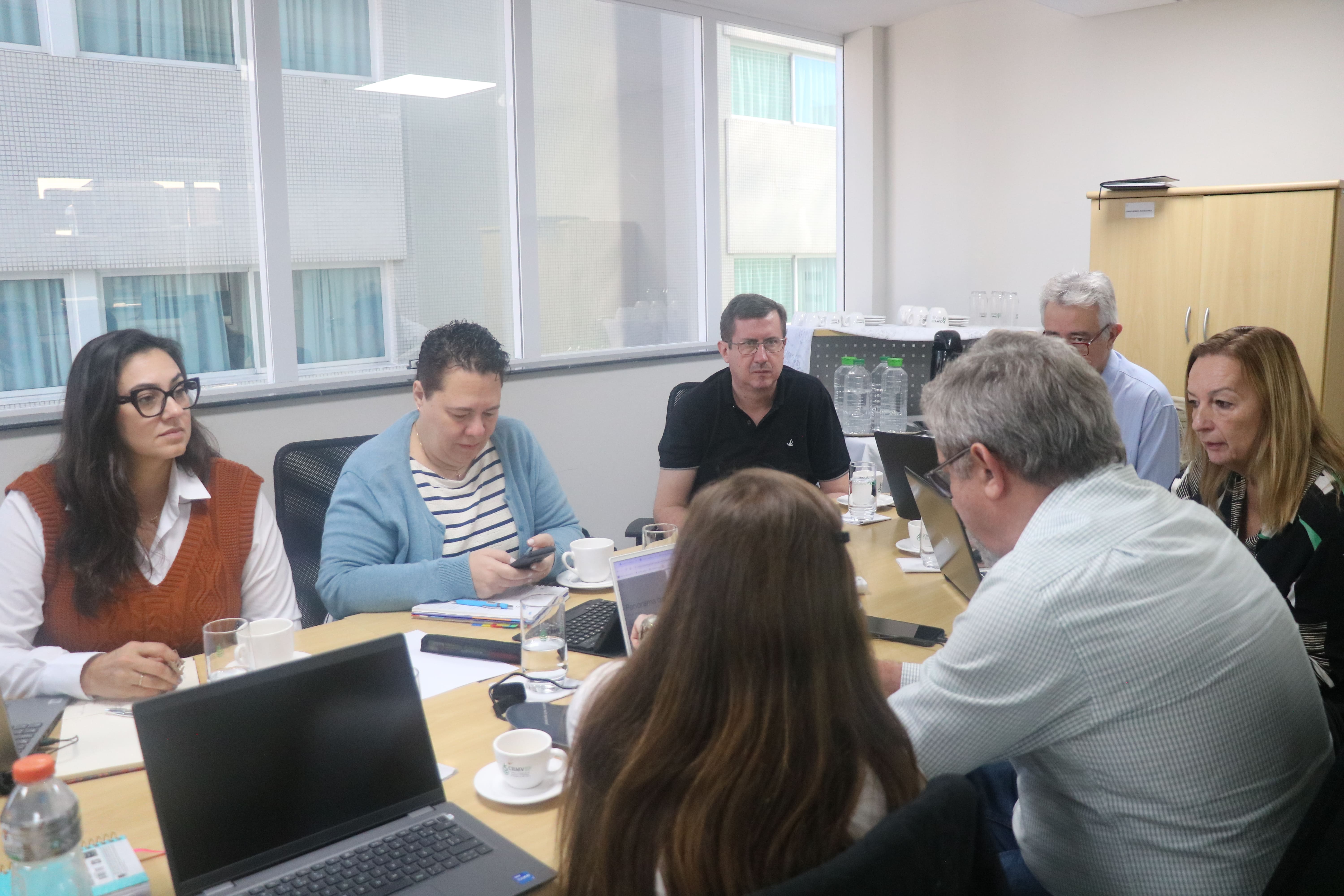 Pessoas reunidas ao redor de uma mesa retangular em reunião.