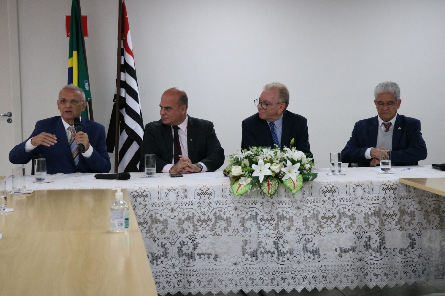 Quatro homens sentados à mesa, um deles fala ao microfone, ao fundo as bandeiras do Brasil e do estado de São Paulo.