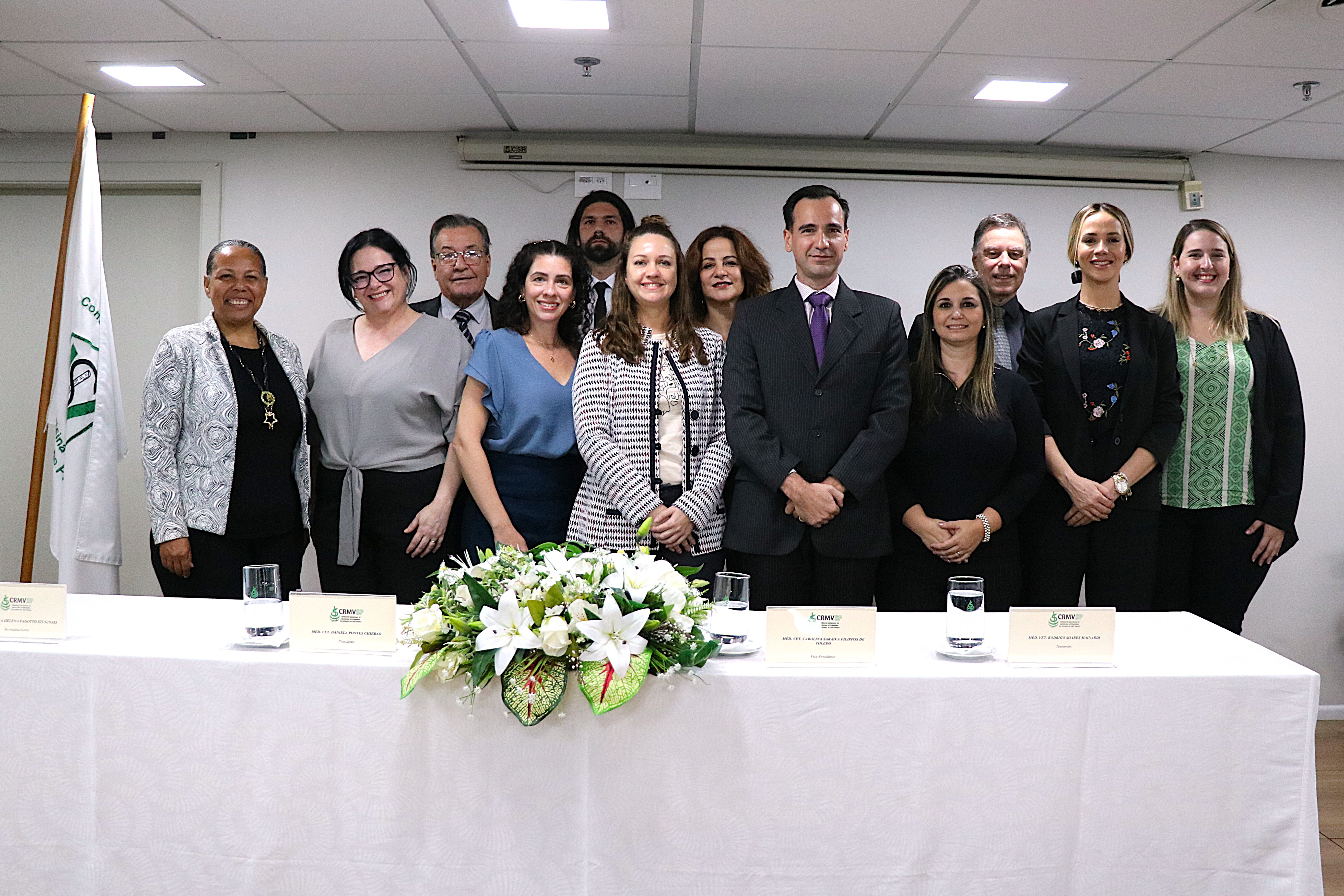 Homens e mulheres em pé posando, com mesa à frente com arranjo de flores. Esses são os conselheiros e diretores do CRMV-SP para o triênio 2024-2027. Ao centro está a presidente do Regional.