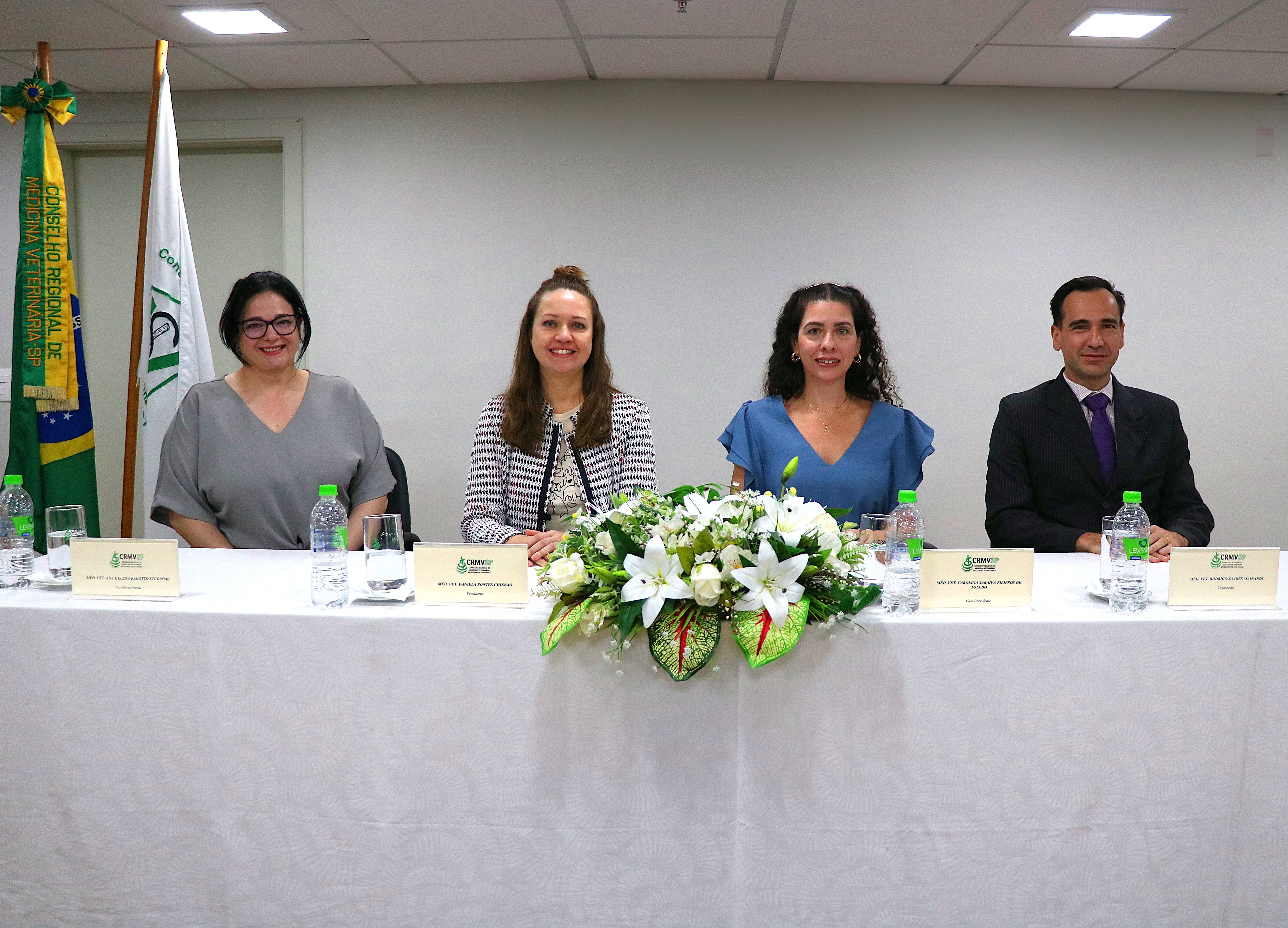 Imagem ilustrativa traz três mulheres e um homem assentados em uma mesa, enfeitada com uma flor no centro, posando e sorrindo. Essa é a nova diretoria executiva do CRMV-SP para o triênio 2024-2027.