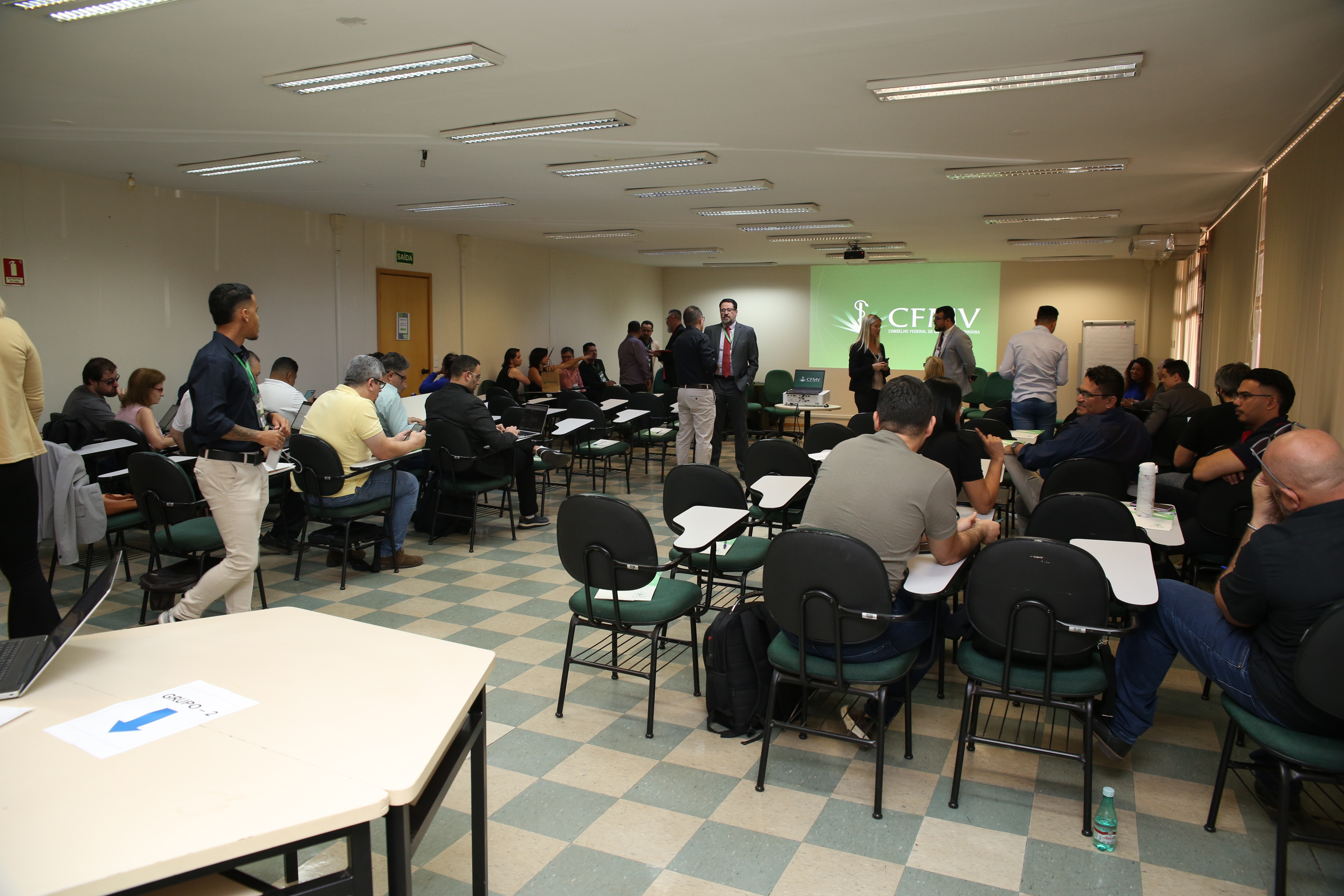 Salão com cadeiras e mesas. À frente, há um telão em que está escrito "CFMV" e o símbolo do Conselho Federal em branco sobre fundo verde. O salão está repleto de pessoas ocupando as cadeiras.