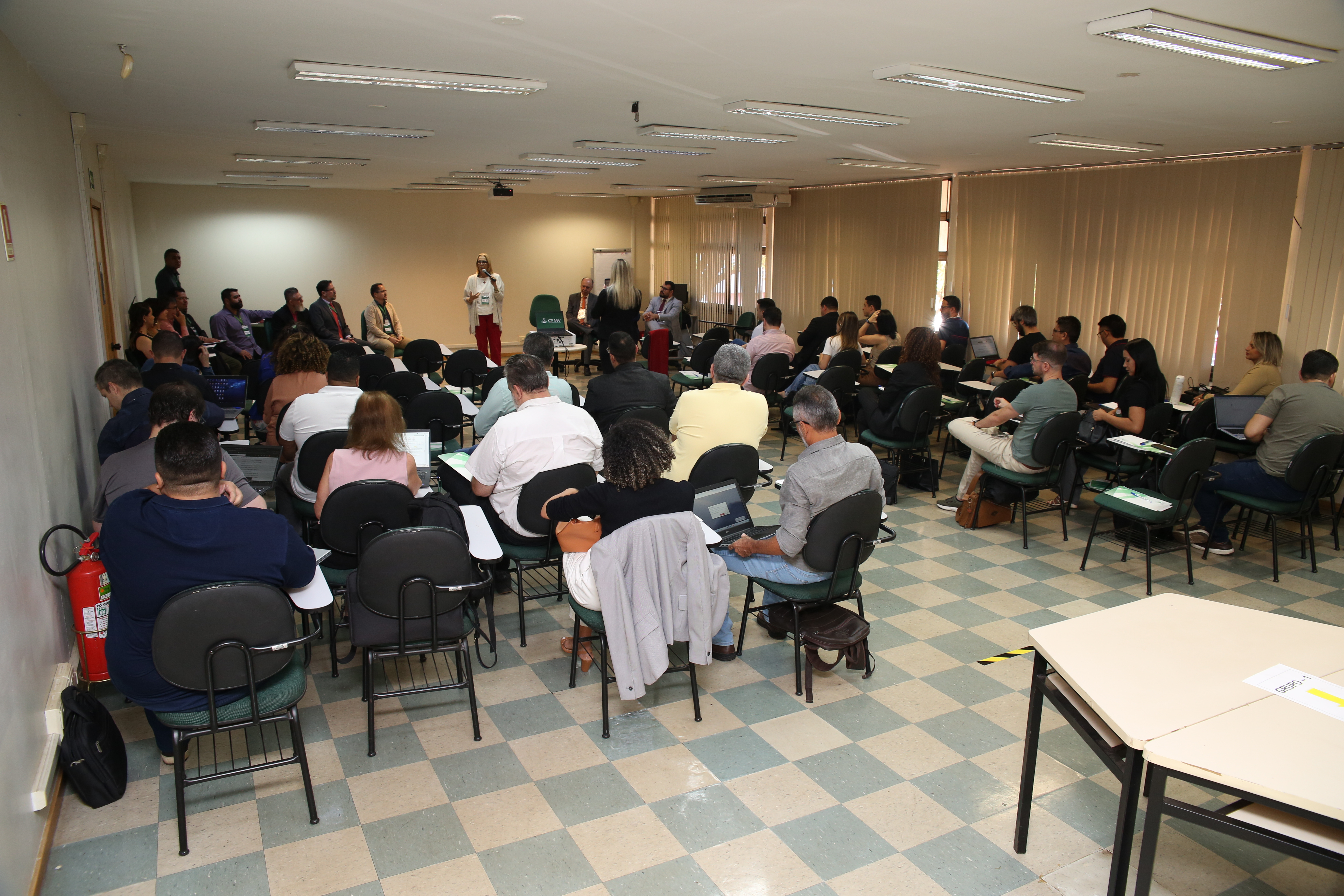 Salão com cadeiras e mesas. O salão está repleto de pessoas ocupando as cadeiras. À frente, está a presidente do CFMV, Ana Elisa Almeida, falando ao microfone.