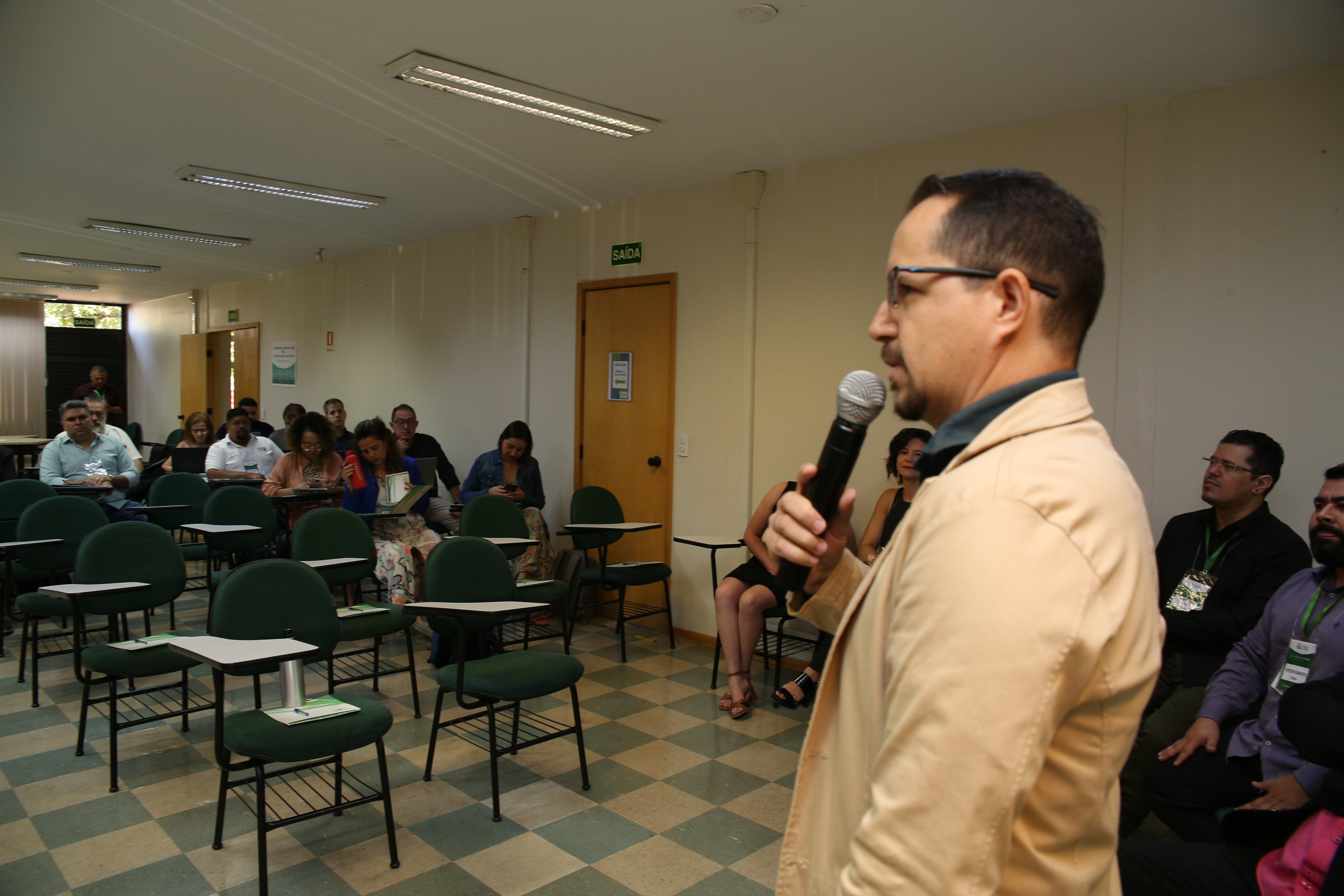 Homem vestindo terno em cor bege está falando ao microfone. Ao fundo, pessoas sentadas em cadeiras estão prestando atenção a ele.
