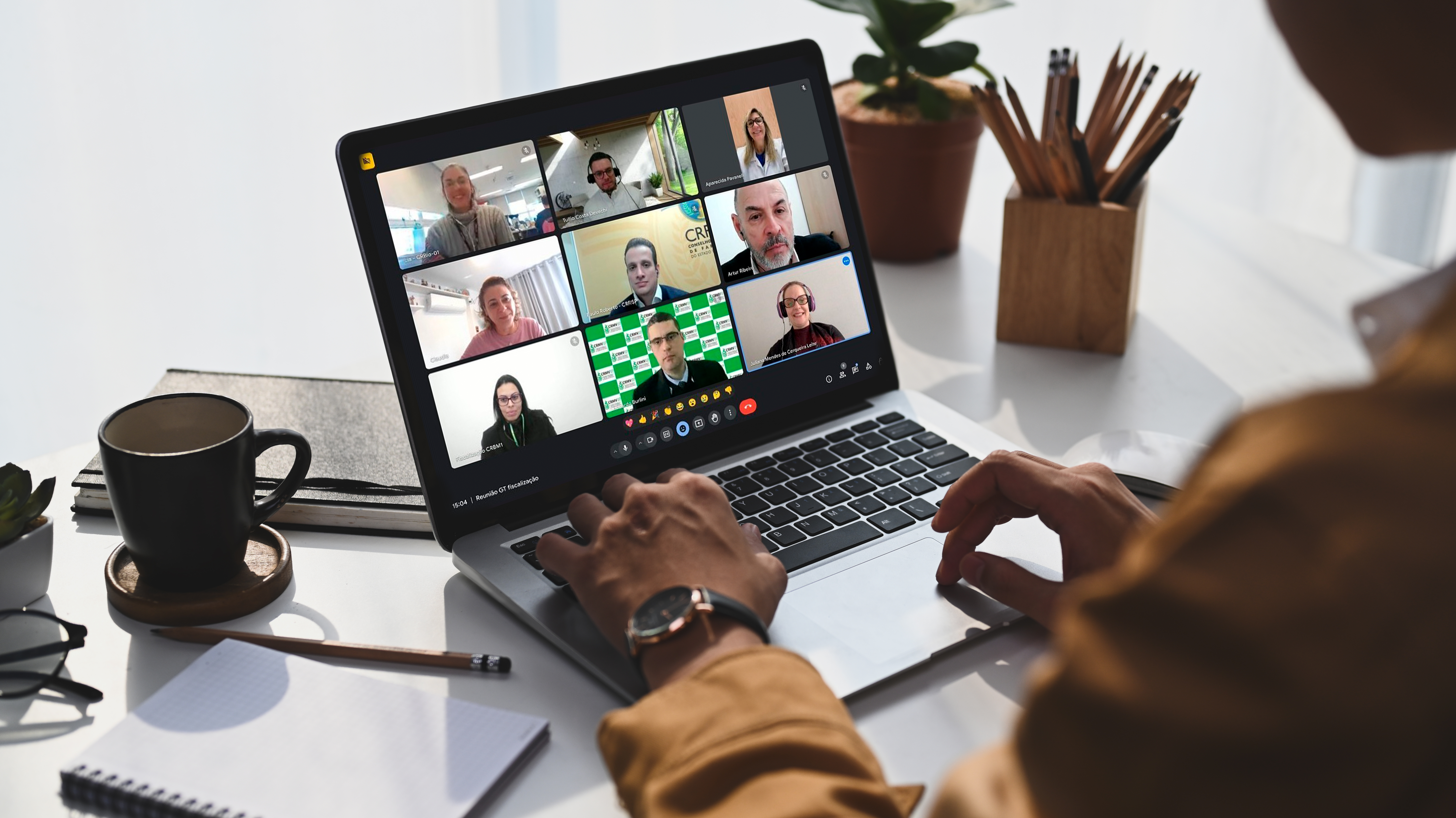 Notebook sobre uma mesa branca em que aparece uma tela de reunião virtual com nove participantes. Sobre a mesa também um óculos, um caerno, uma xícara, alguns lápis e uma planta. Uma pessoa também mexe no equipamento, mas na imagem aparece apenas duas mãos sobre o teclado.