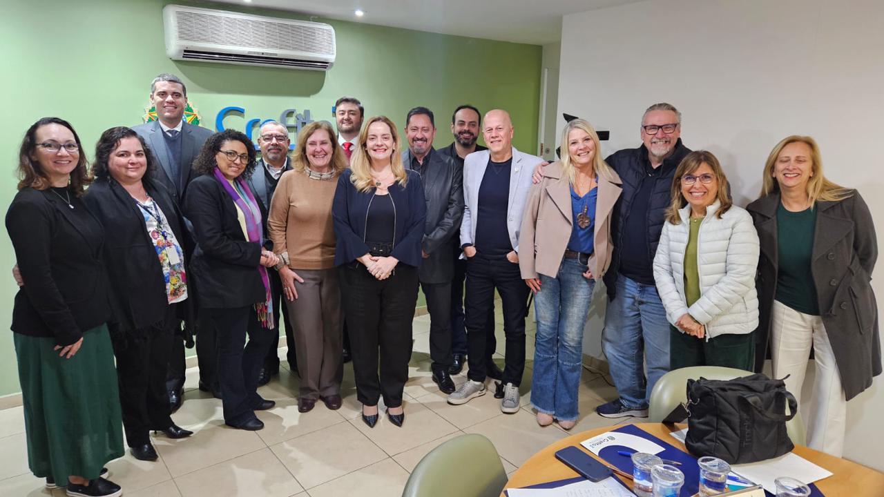 Representantes de 12 conselhos profissionais estão reunidos de pé e sorrindo para a foto. Ao todo são 15 pessoas, sendo oito mulheres e sete homens. Ao centro, a coordenadora do Fcafs.
