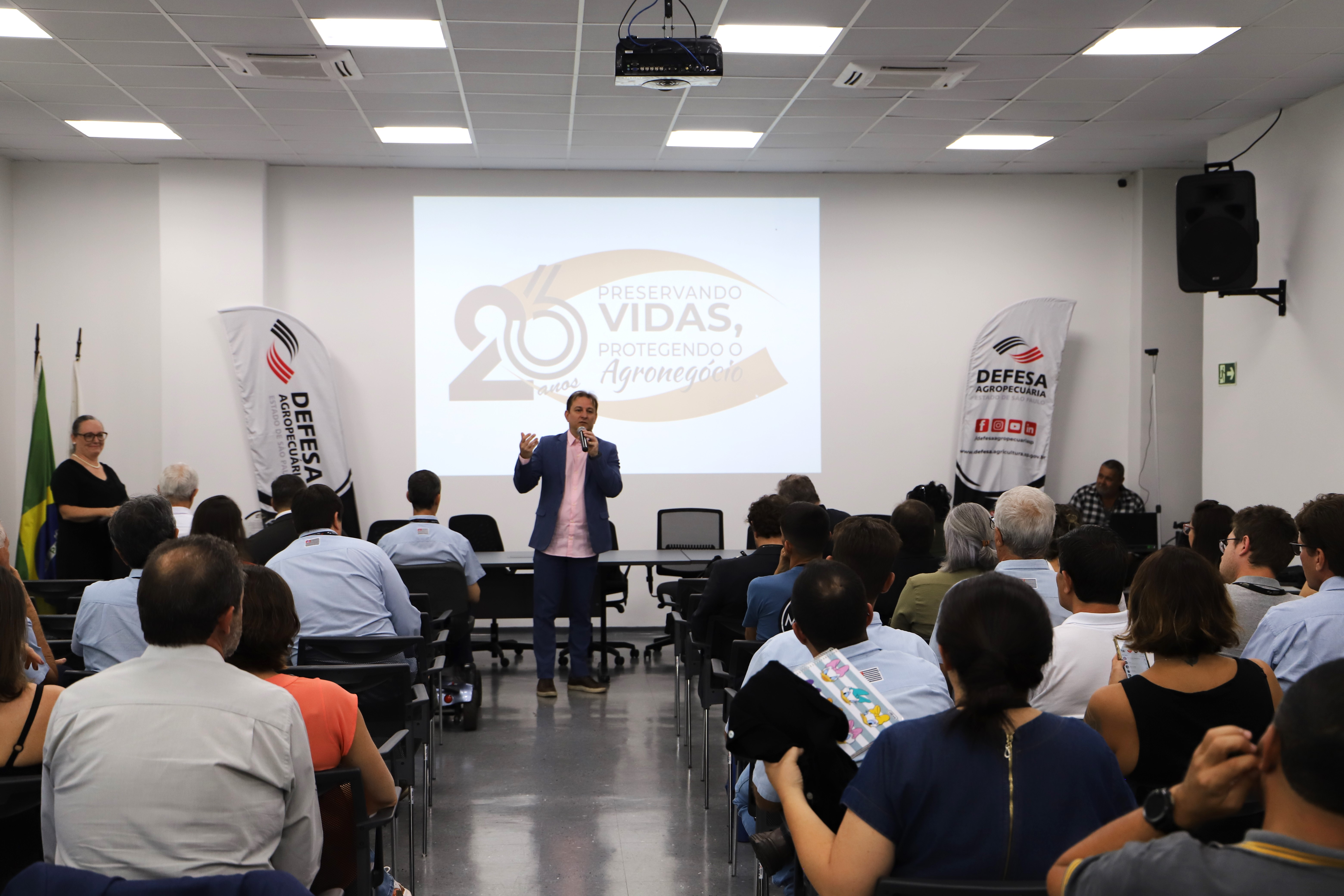 Homem discursando enquanto pessoas assistem sentadas em um auditório. No telão, a mensagem: 26 anos - Preservando vidas, protegendo o agronegócio.