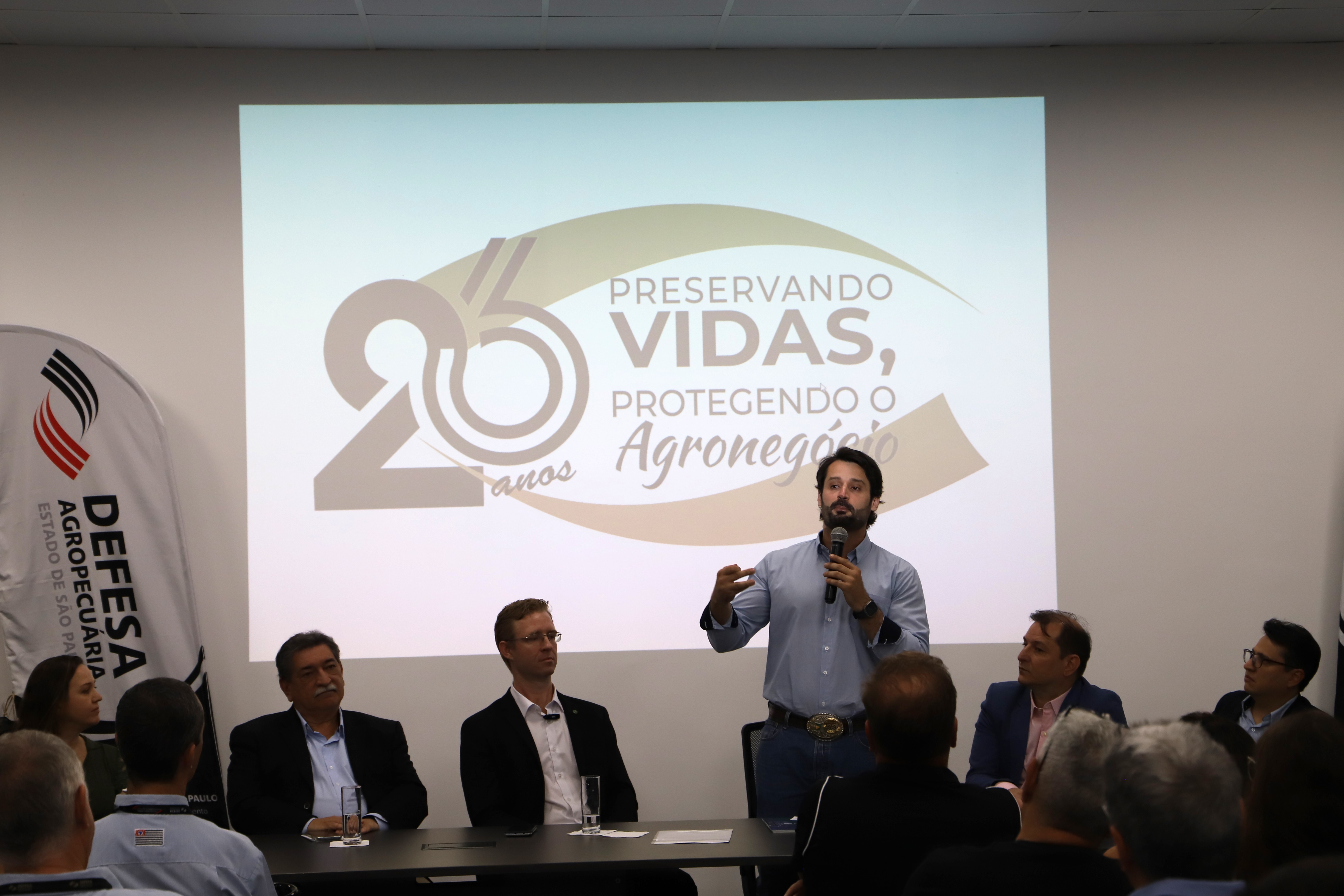 Homem discursando de pé, enquanto pessoas seguem sentadas na mesa de autoridades, e outras assistem sentadas na plateia.