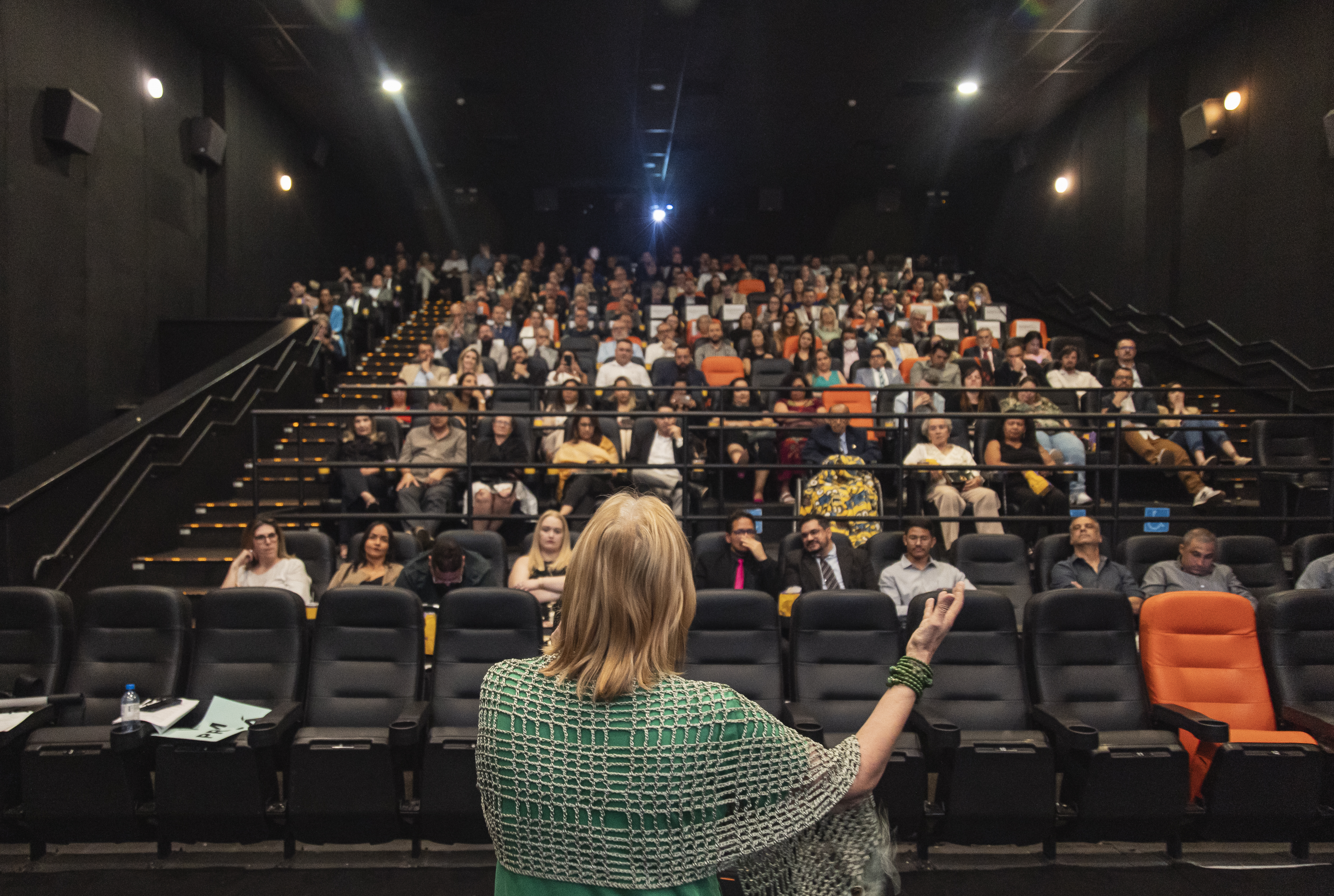 Uma sala de cinema com varias pessoas sentadas ouvindo e uma mulher de costas para a foto discursando para o público.