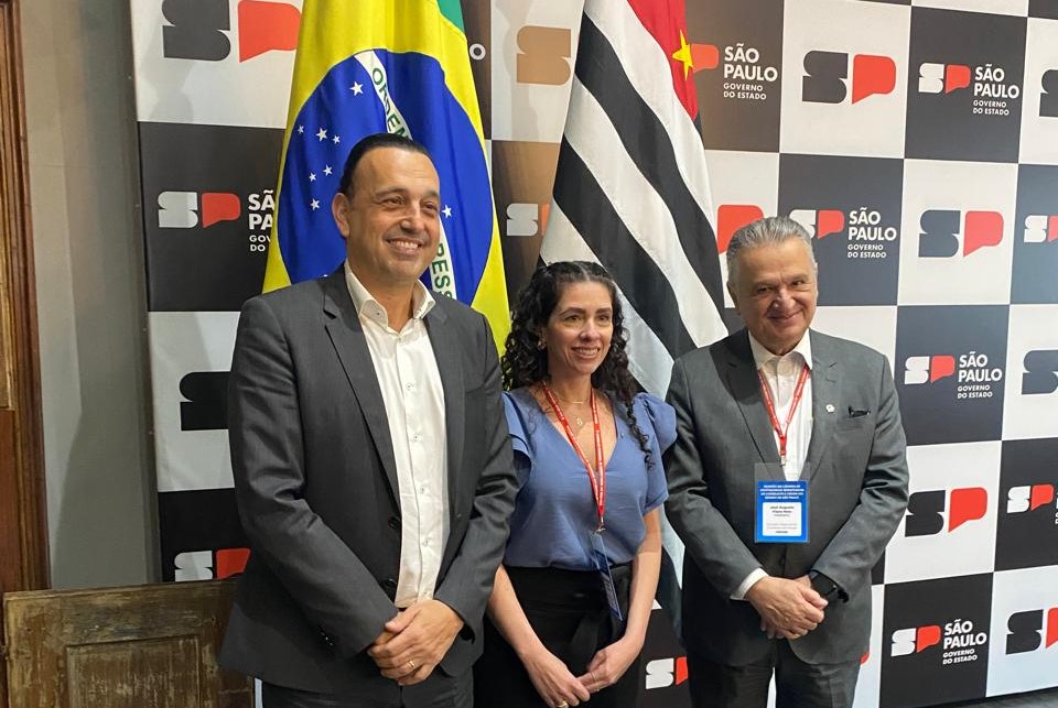 Dois homens e uma mulher (centro) posam para a foto. Ao fundo, painel com a identificação do estado de São Paulo, com as bandeiras do Brasil e de São Paulo.