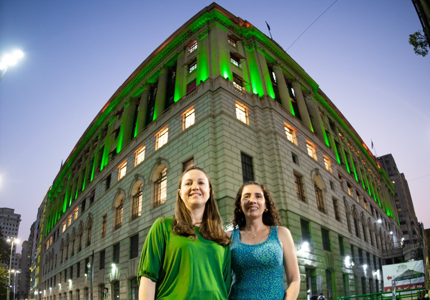 Em primeiro plano, duas mulheres sorrindo. Atrás edifício iluminado nas cores verdes.