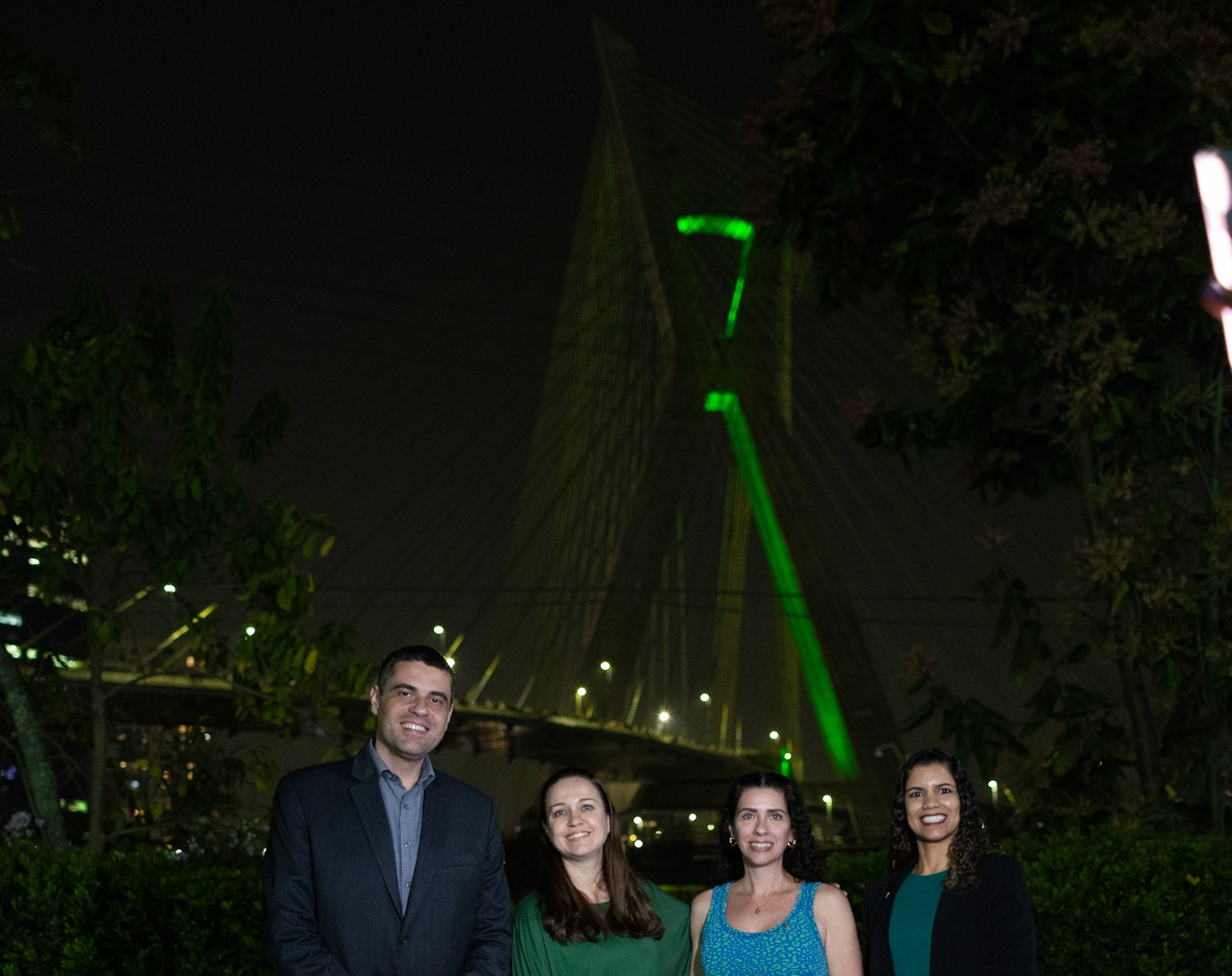 Um homem e três mulheres em primeiro plano, atrás ponte estaiada iluminada em verde.