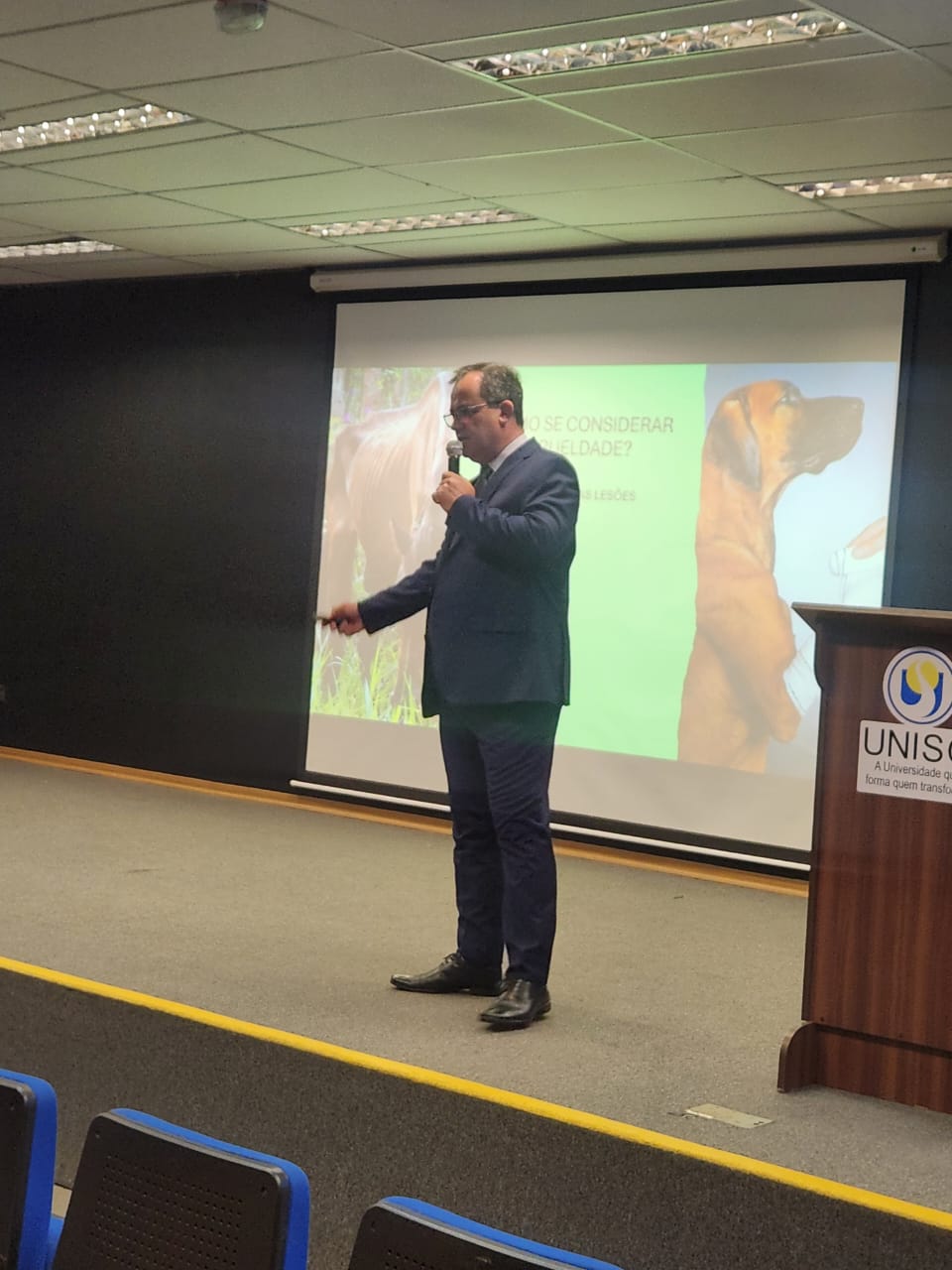 Homem no palco em frente ao telão palestrando.