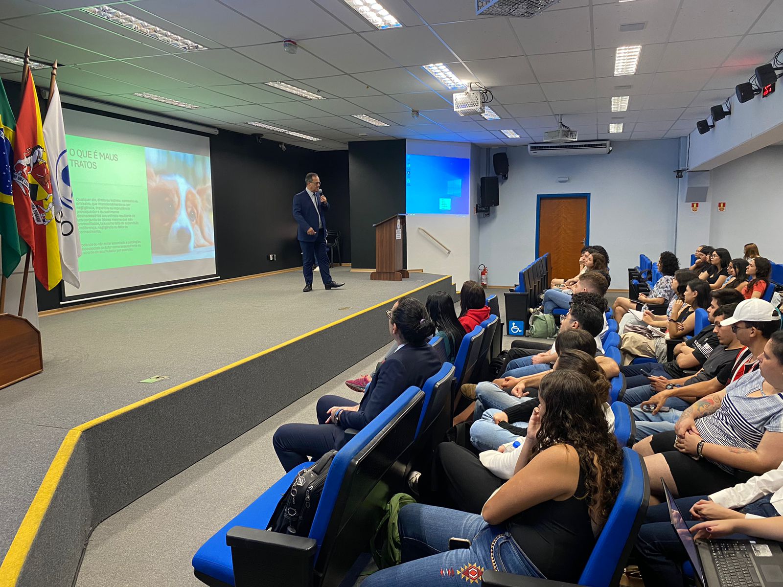 Homem está no palco em frente a um telão diante de auditório lotado de pessoas.