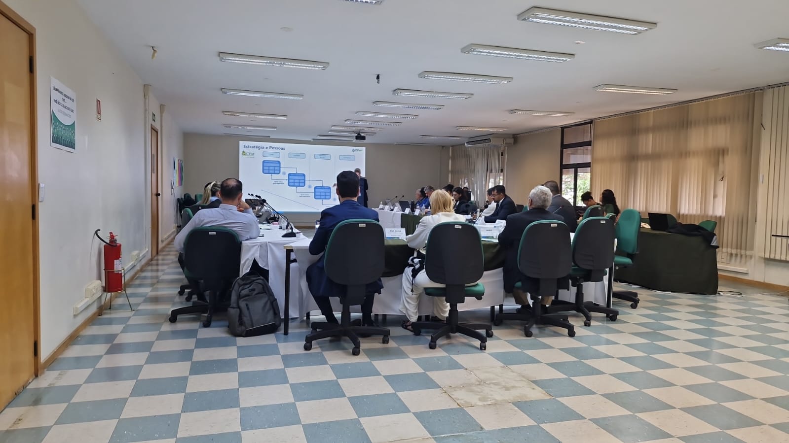 Pessoas sentadas em reunião de costas viradas para a câmera.
