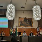 Pessoas juntas sentadas em cadeiras beges falando na mesa principal, ao fundo, parede metade de madeira e metade superior azulejada com um telão mostrando uma planilha e na direita, quadro de moldura dourada retratando o período colonial brasileiro. Acima da mesa dois lustres de cristal em formato redondo.