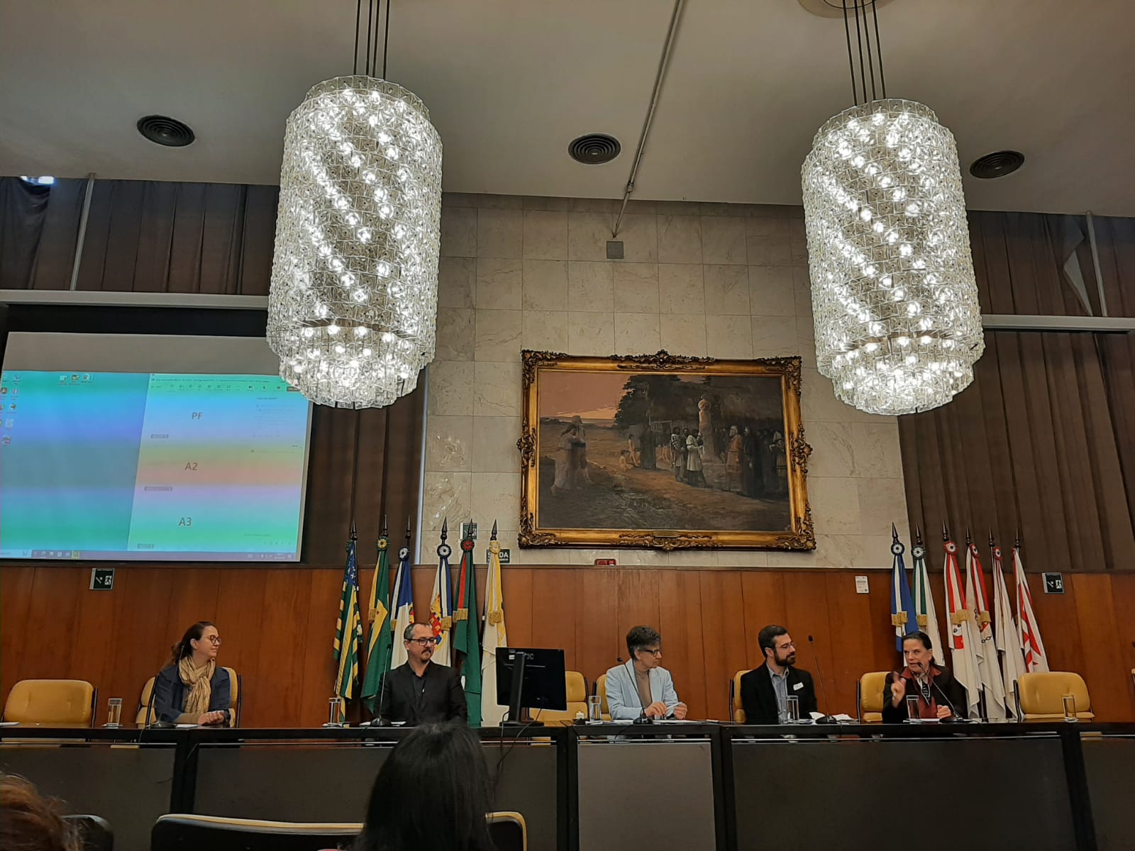 Pessoas juntas sentadas em cadeiras beges falando na mesa principal, ao fundo, parede metade de madeira e metade superior azulejada com um telão mostrando uma planilha e na direita, quadro de moldura dourada retratando o período colonial brasileiro. Acima da mesa dois lustres de cristal em formato redondo.