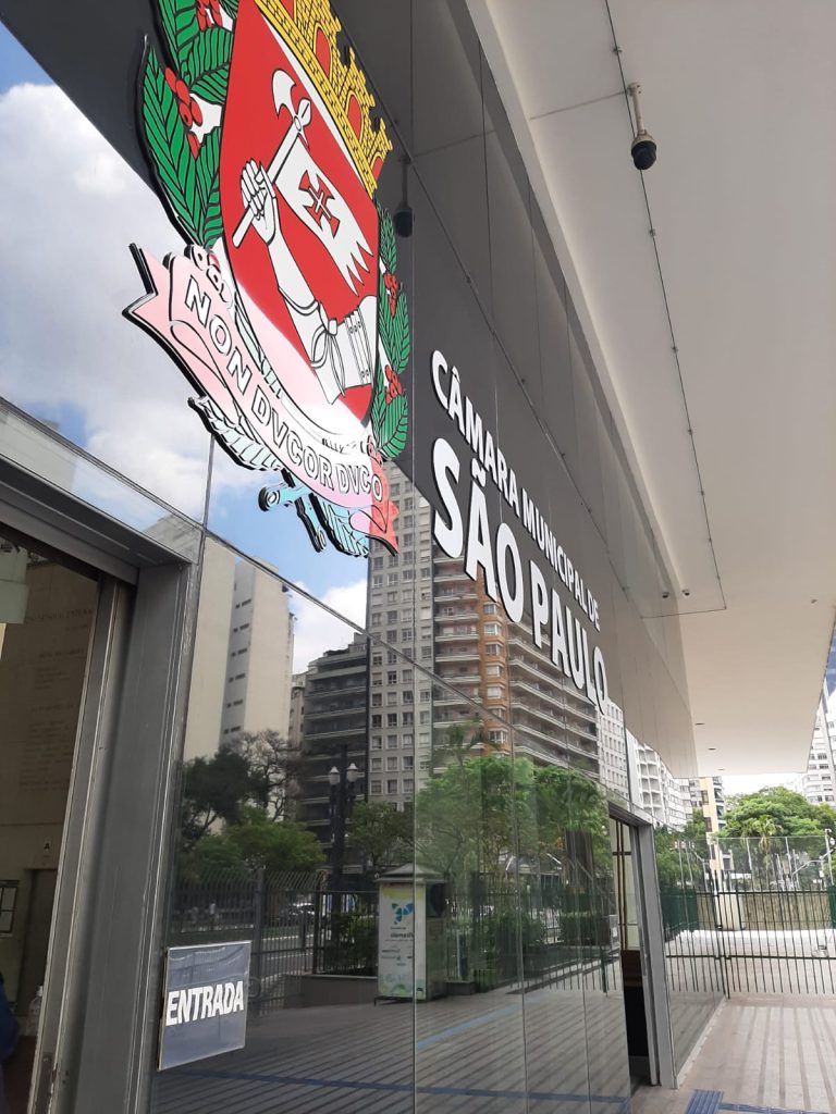 Fachada da Câmara Municipal de São Paulo, que é composta de vidros pretos espelhados com foco para o escudo da cidade de São Paulo e os dizeres "Câmara Municipal de São Paulo."