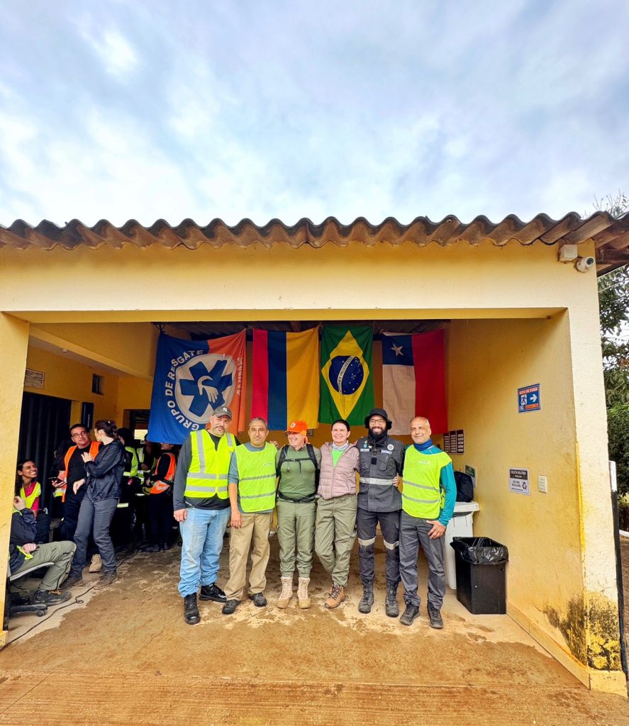 Pessoas juntas posando para foto, atrás delas uma bandeira do grupo de resgate, a bandeira da Armênia, a do Brasil e a do Chile.