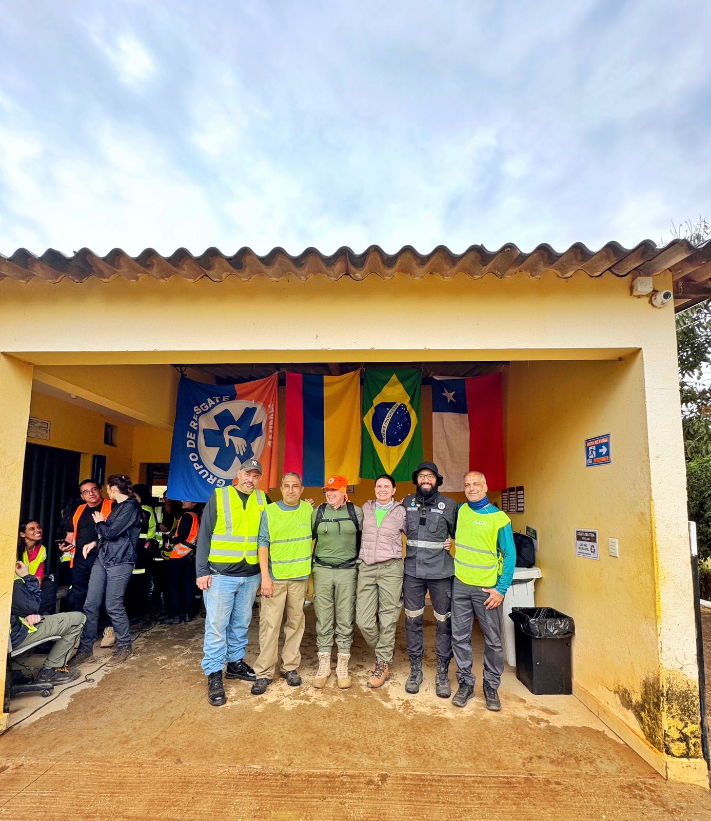 Pessoas juntas posando para foto, atrás delas uma bandeira do grupo de resgate, a bandeira da Armênia, a do Brasil e a do Chile.