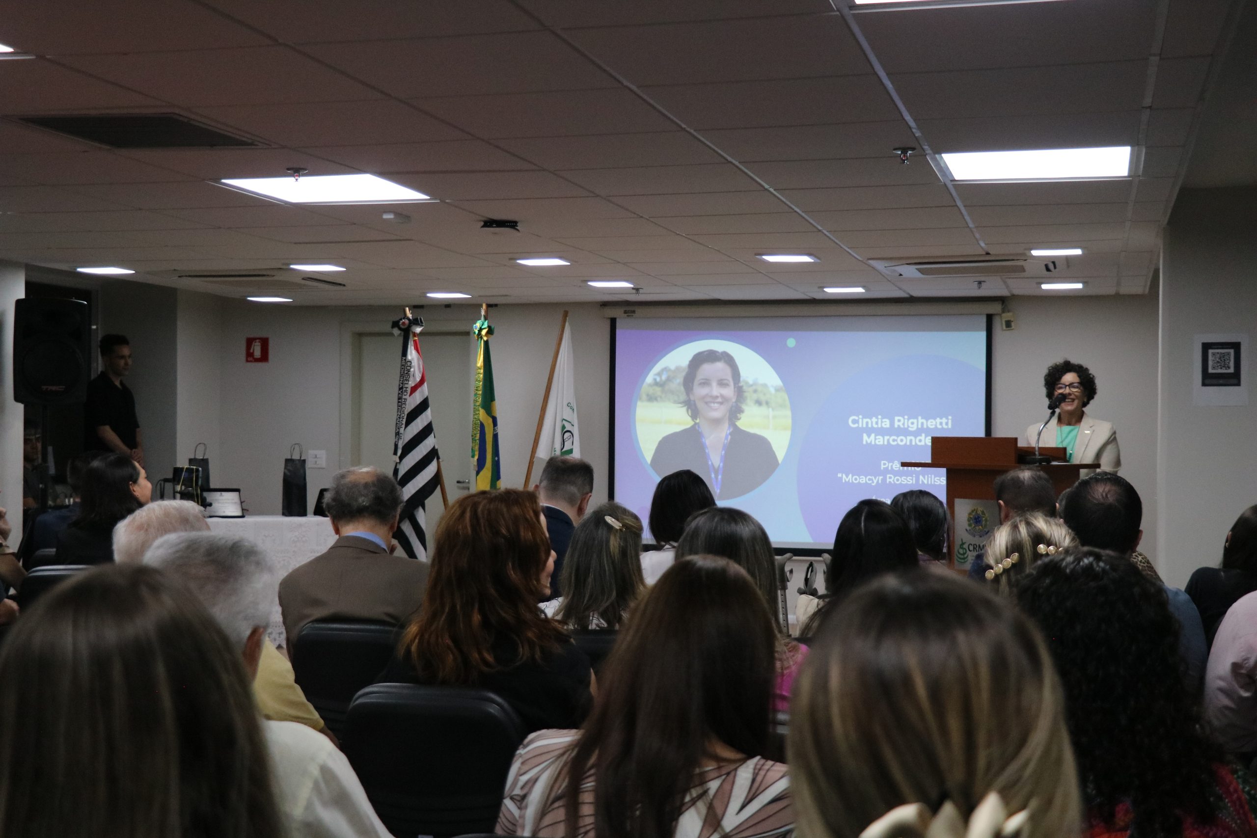 A premiada na categoria Pesquisa discursa no púlpito para auditório lotado, ao fundo sua foto aparece no telão.