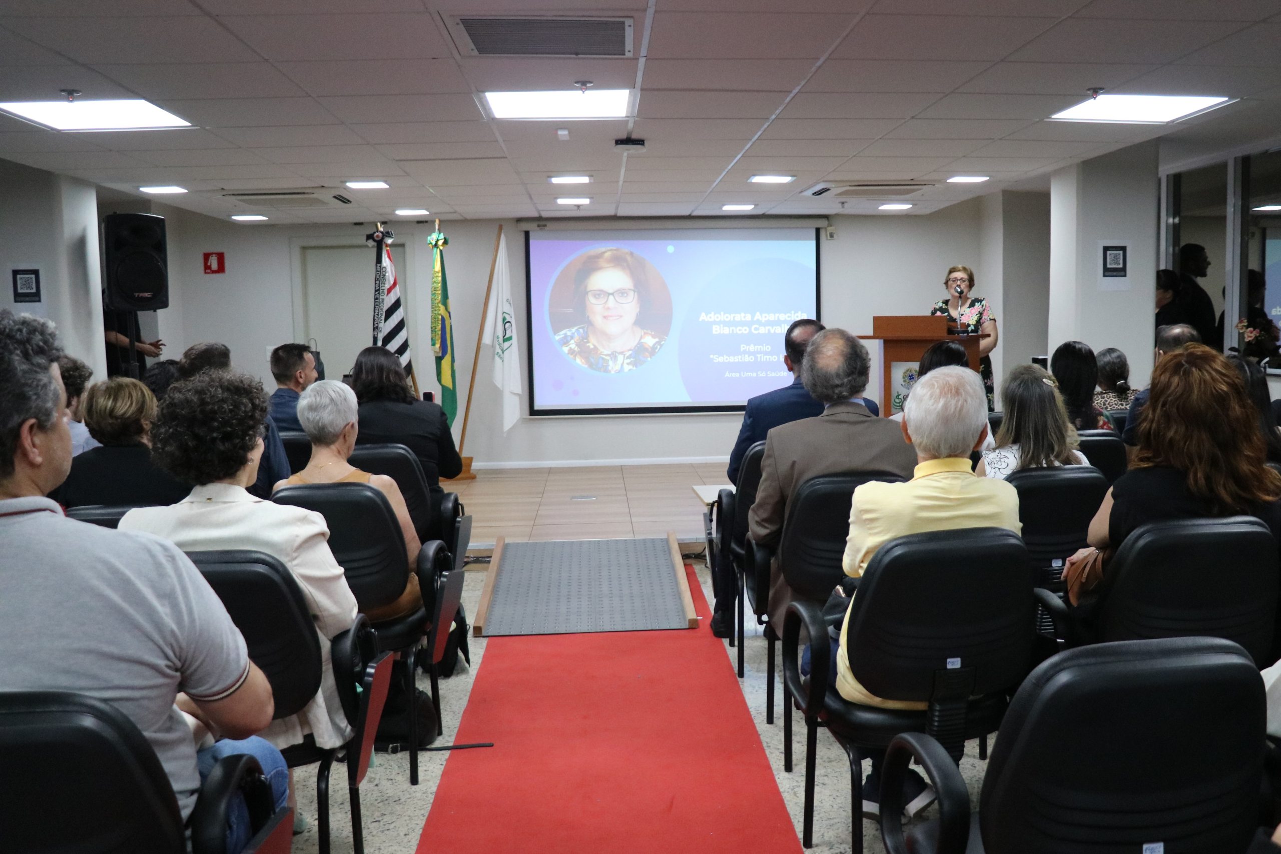 A premiada na categoria Uma Só Saúde discursa para o auditório lotado, ao fundo uma foto sua aparece no telão.